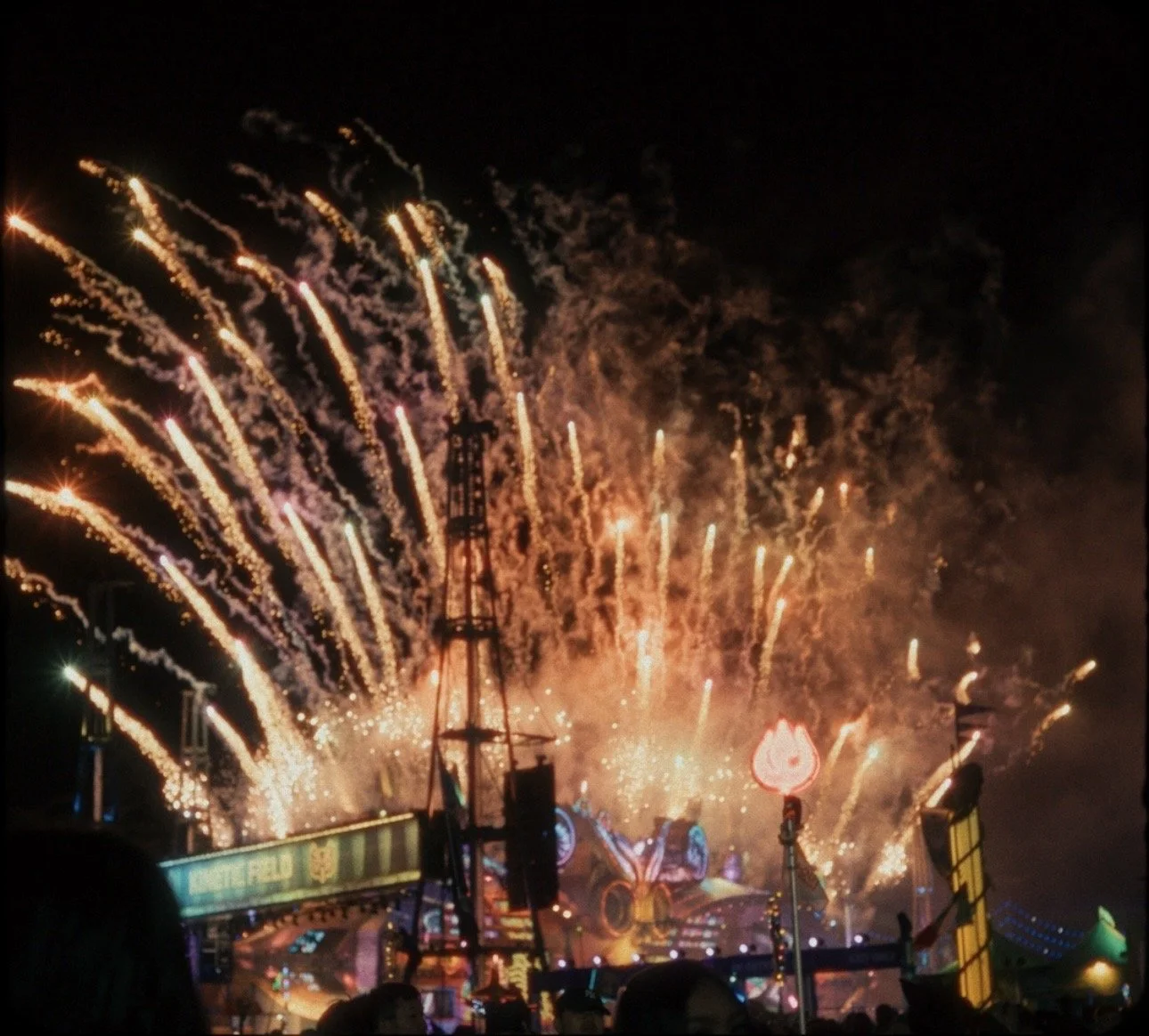 Nighttime fireworks display over a festival or carnival with colorful explosions in the sky, illuminated rides and structures in the foreground, and silhouettes of people watching.
