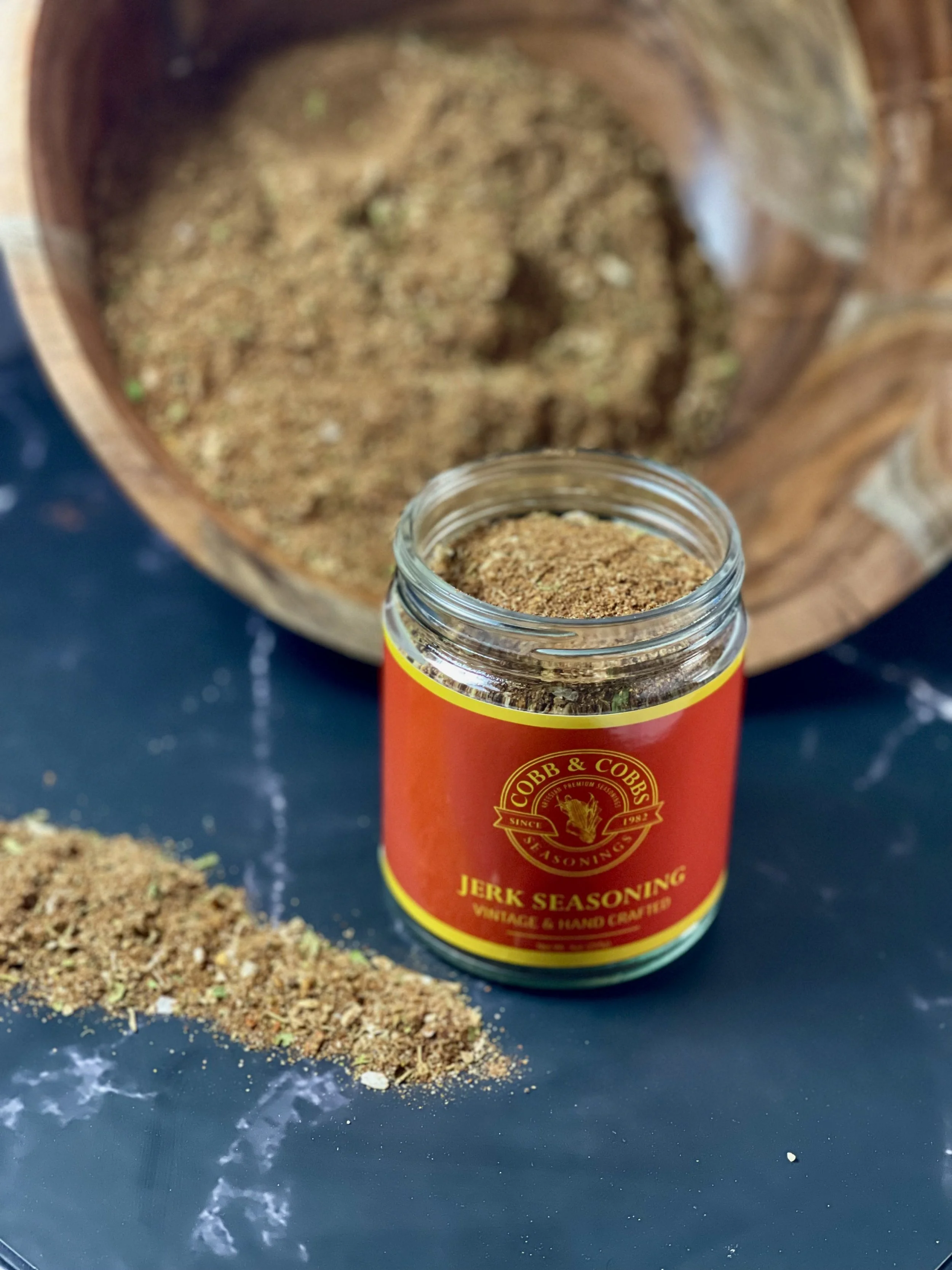 Jar of jerk seasoning on a black surface with spilled seasoning and a wooden bowl in the background.