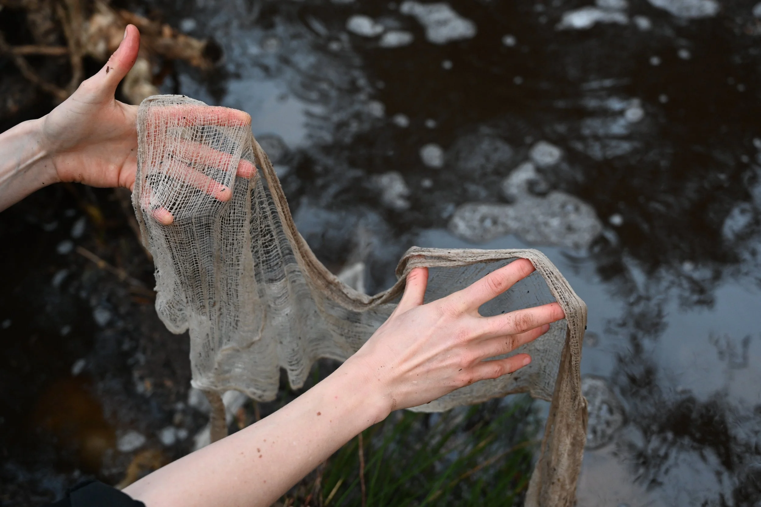 Two hands holding a weathered, translucent fabric or netting near a body of water with dark, bubbling water and rocks in the background. Copyright Kate Alida Mullen