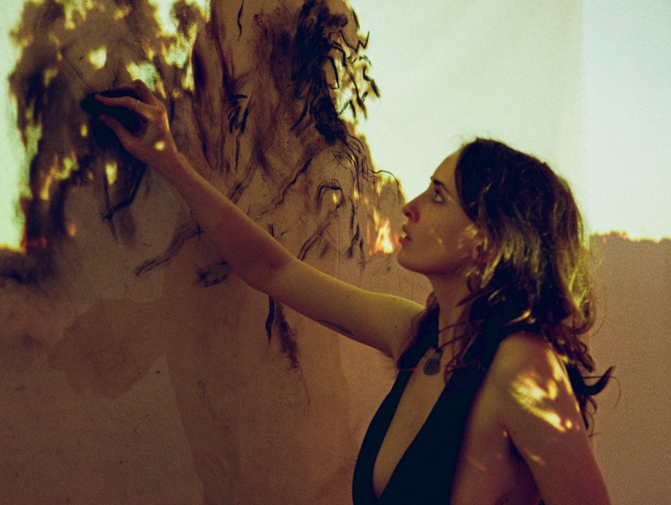 A woman with long, curly hair is touching the wall with her hand, looking at a shadowy figure of a man with long hair and a beard painted on the wall. The lighting is dim and warm. Copyright Kate Alida Mullen