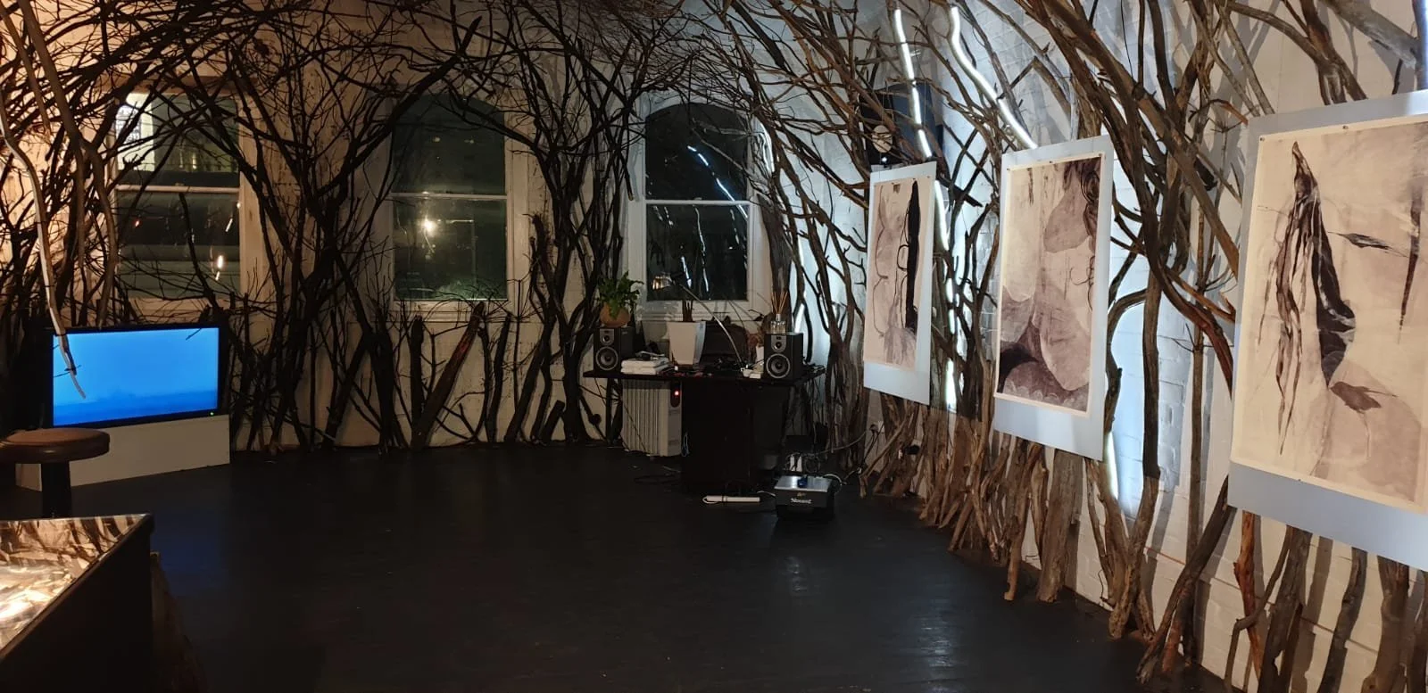 An art exhibition space with walls decorated with intertwined branches, featuring several framed artworks hung in a row. There is a small table or booth with audio equipment, and a television or monitor on the left side of the room displaying a blue 