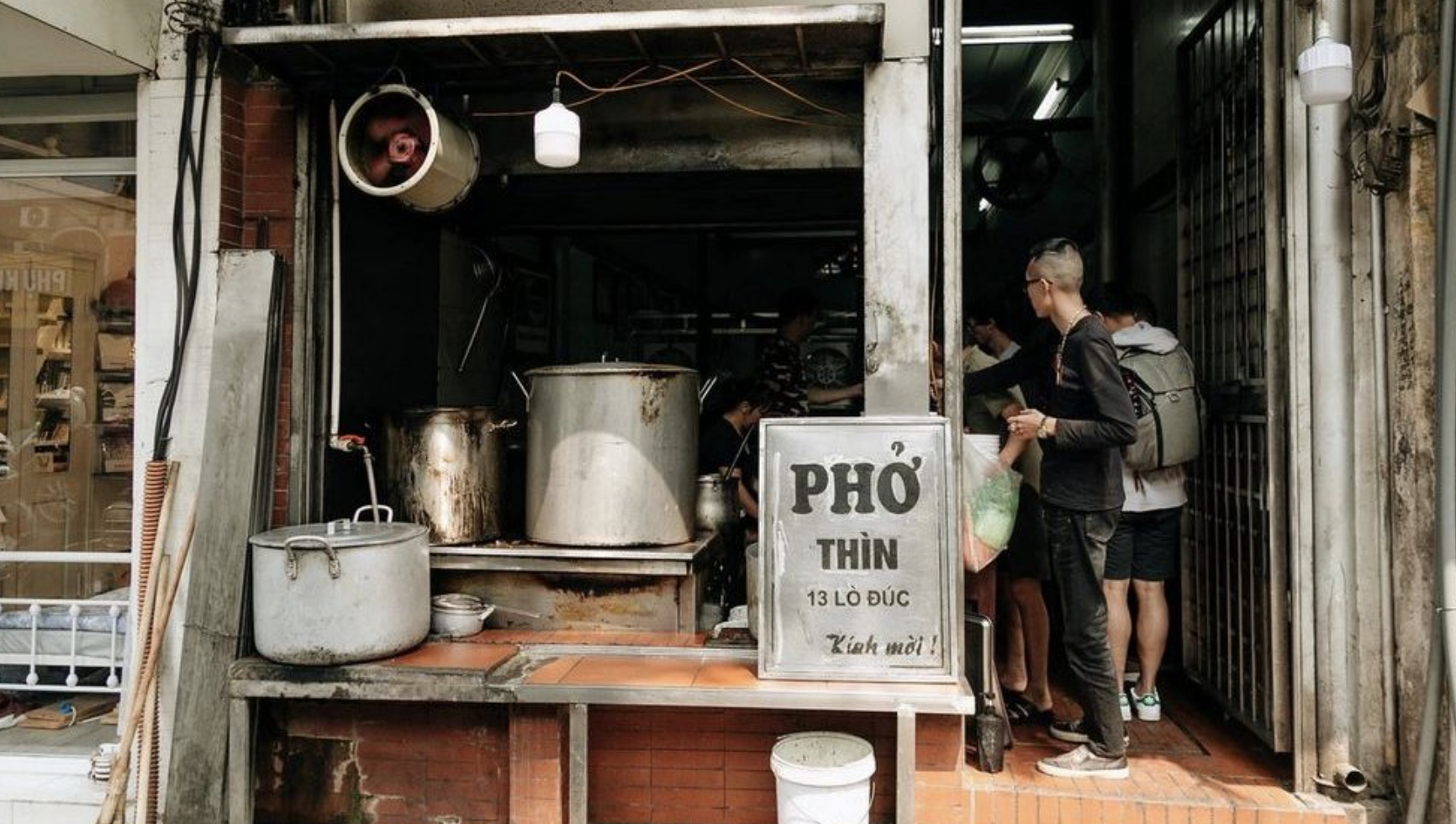 Street food stall with large cooking pots, a sign with Vietnamese writing, and customers ordering food.