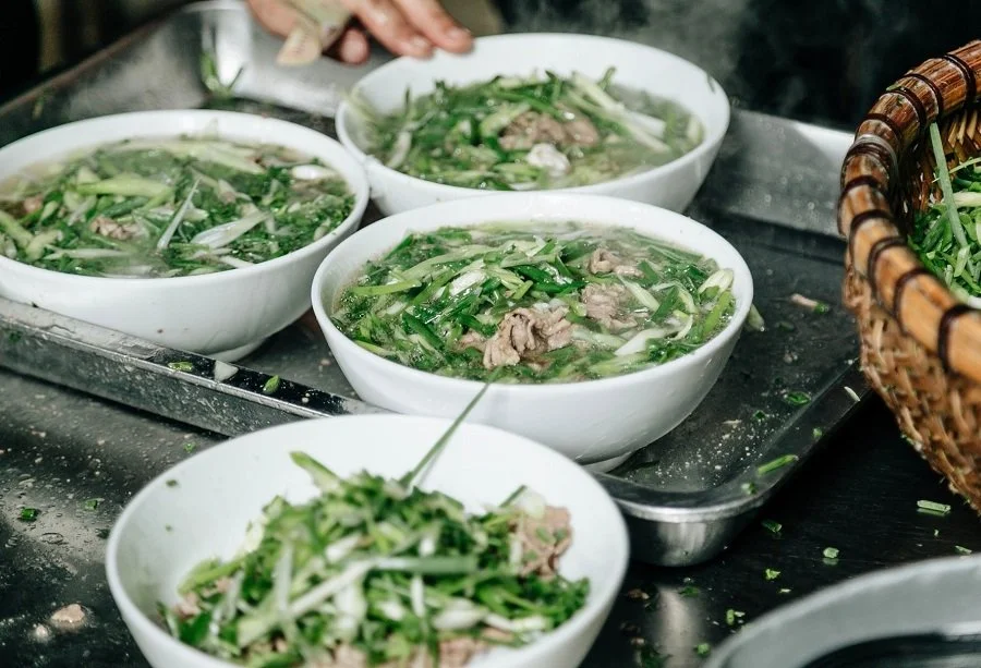 Four bowls of hot Vietnamese Pho soup with green onions, herbs, and slices of cooked meat on a metal tray, with chopsticks, herbs, and a wicker basket nearby.
