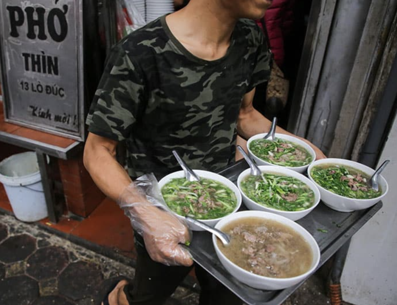 A man in a camouflage T-shirt holding a tray with five bowls of pho, a Vietnamese noodle soup, in an outdoor street setting.