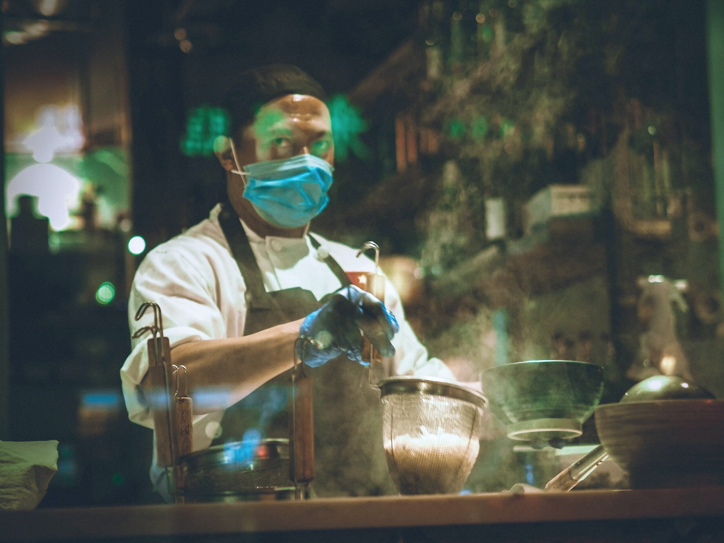 A chef wearing a face mask and gloves preparing food in a kitchen at night, seen through a glass window.