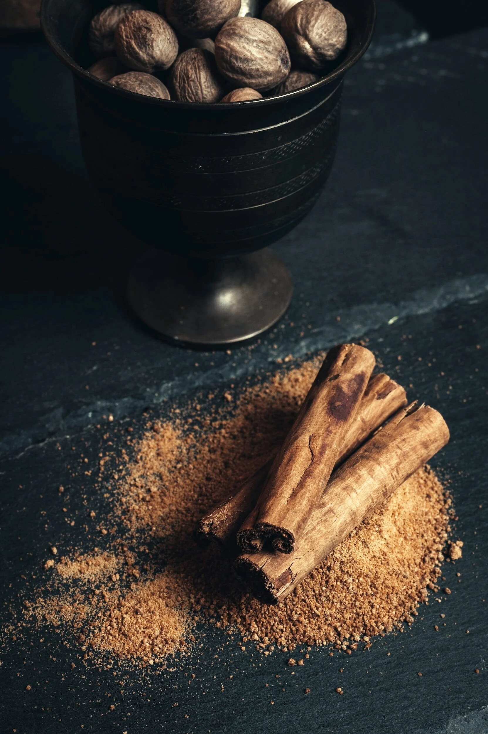 A black cup filled with whole nutmeg seeds, a few cinnamon sticks resting on a dark surface with ground cinnamon sprinkled around.