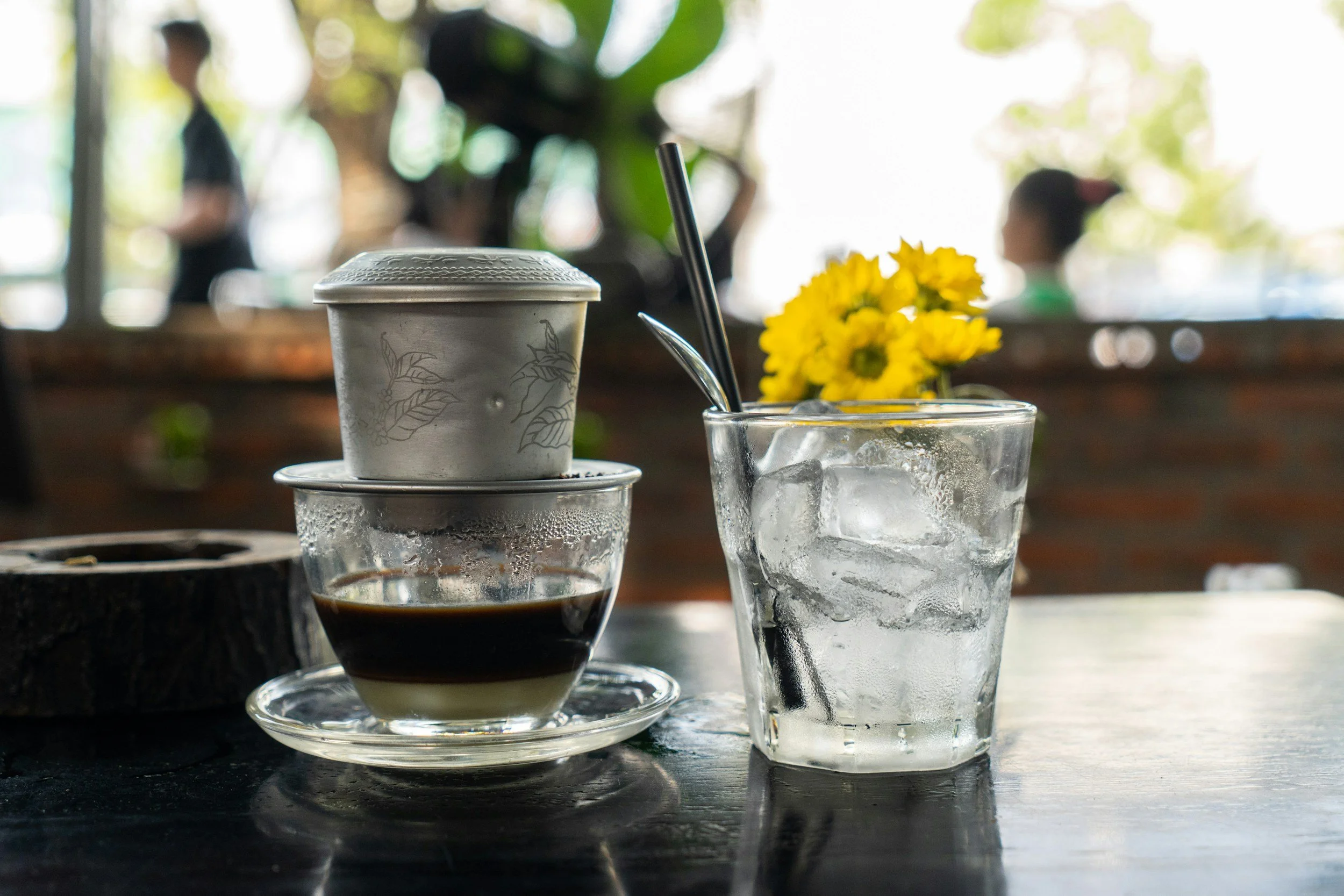 A glass of iced water, a glass cup of Vietnamese black coffee with a lid on a saucer, a small container with a spoon, and a yellow flower in the background on a table inside a restaurant.