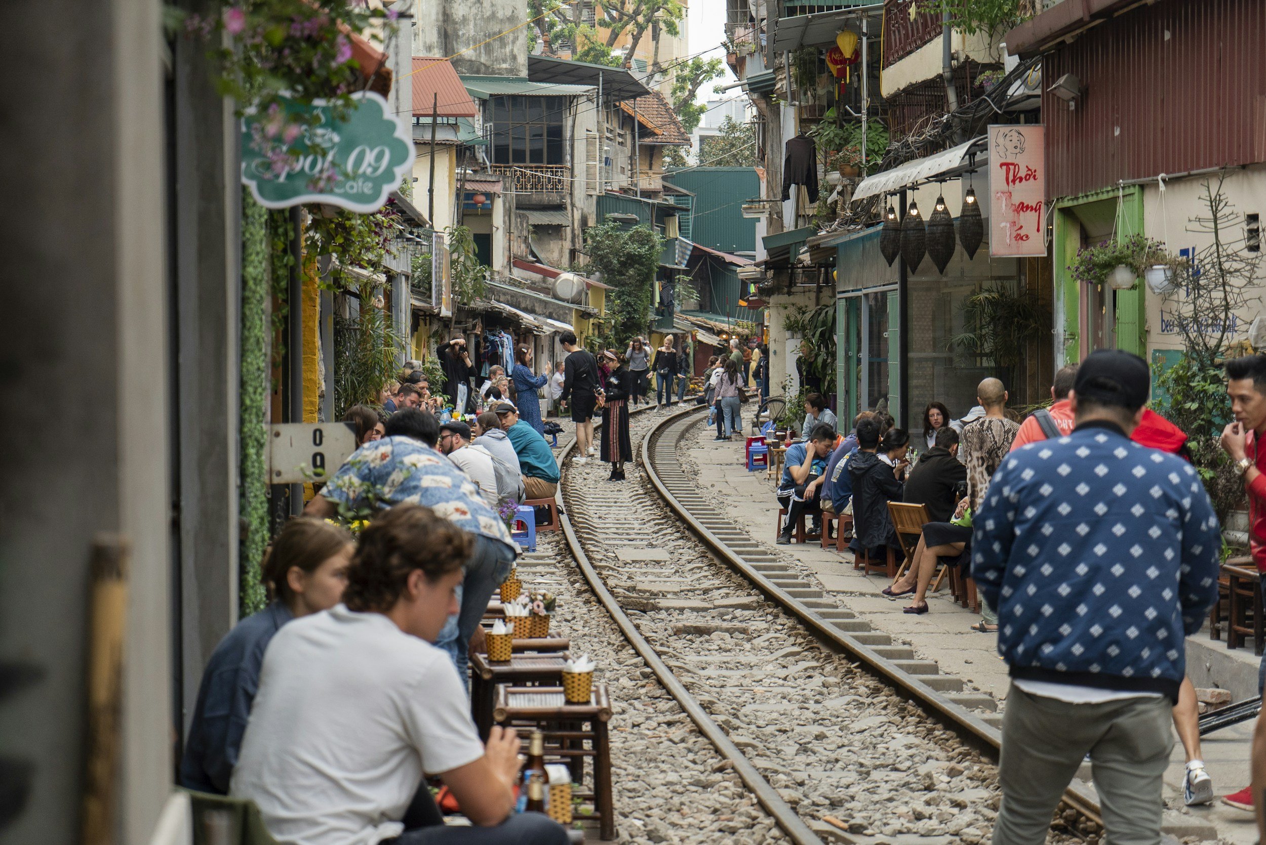 People sitting and eating along a street with Hanoi train tracks, crowded with pedestrians and shops, in an urban area.
