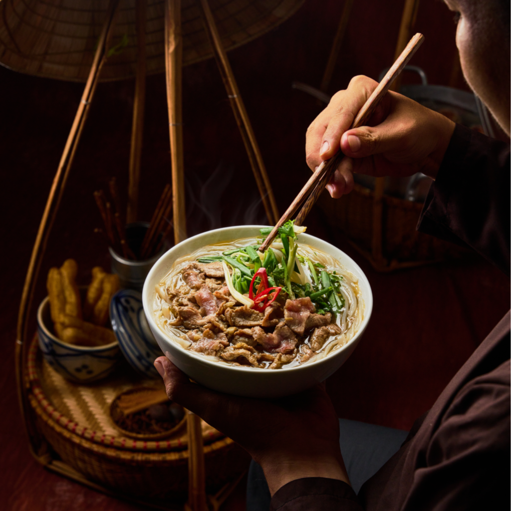 A person holding a bowl of Asian Pho beef noodle soup with sliced meat and green vegetables, using chopsticks. In the background, there is a wicker umbrella, a small dish of fried items, and a container with chopsticks on a woven tray.