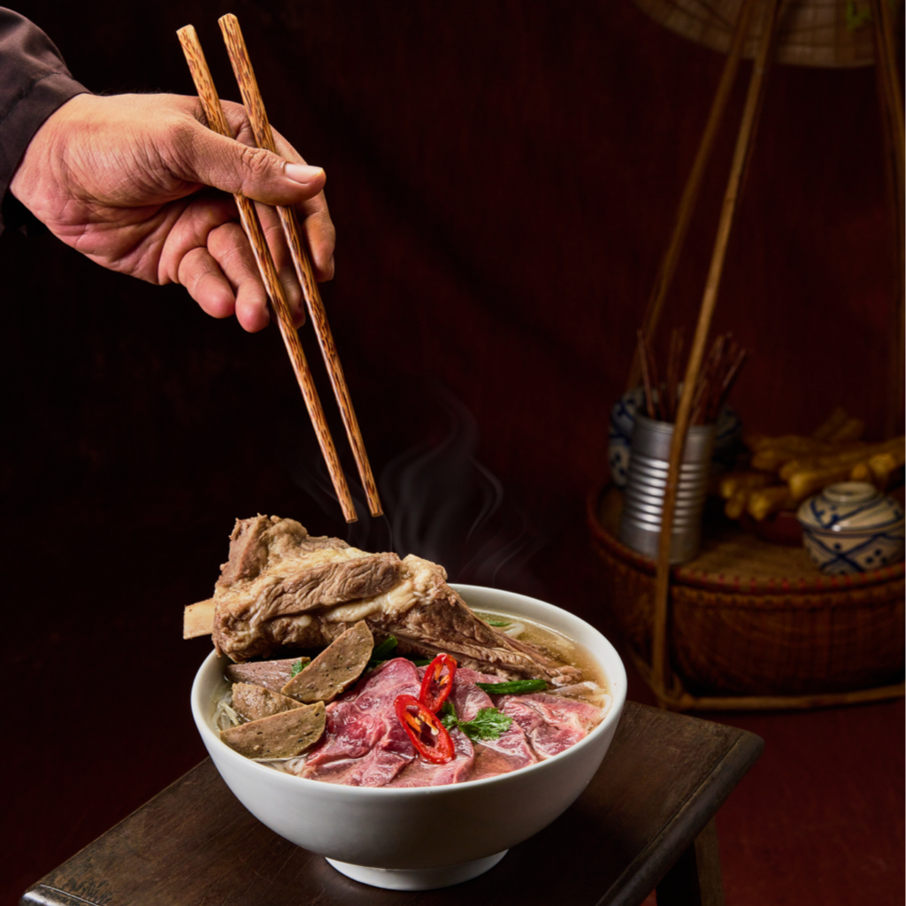 A hand using chopsticks to pick up sliced beef and ribeye and herbs from a steaming bowl of hot Pho beef noodle soup.