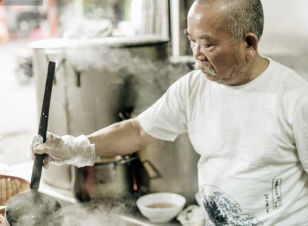 An elderly man with a white t-shirt and gray hair cooking in a kitchen, stirring a pot on a stove.