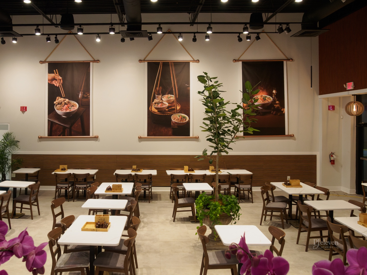 Dining area in a restaurant with white tables, wooden chairs, a large potted plant in the center, and decorative wall hangings of Asian dishes.