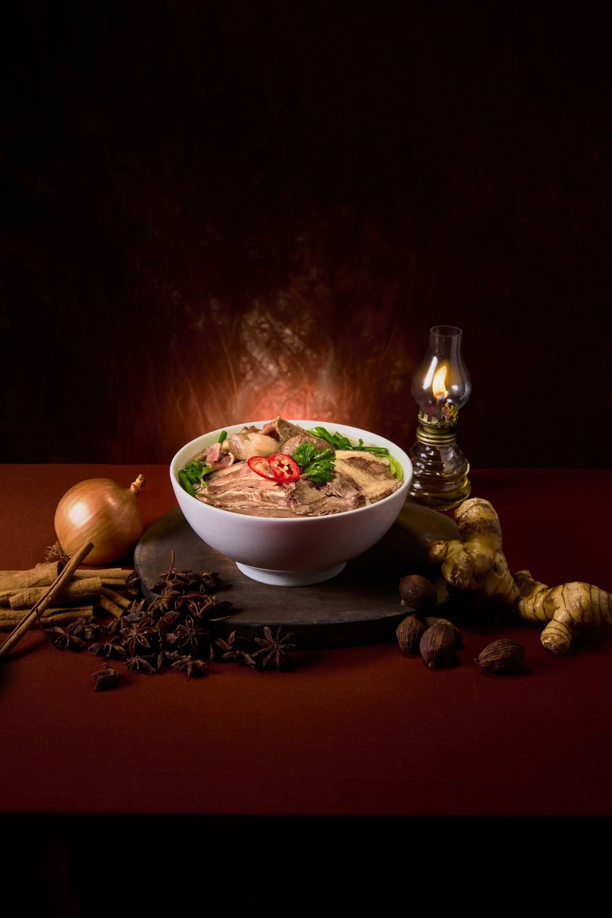 A bowl of steaming hot Vietnamese pho surrounded by herbs and spices, including ginger, star anise, black peppercorns, onions, and ginger roots, with a lit oil lamp beside it against a dark background.