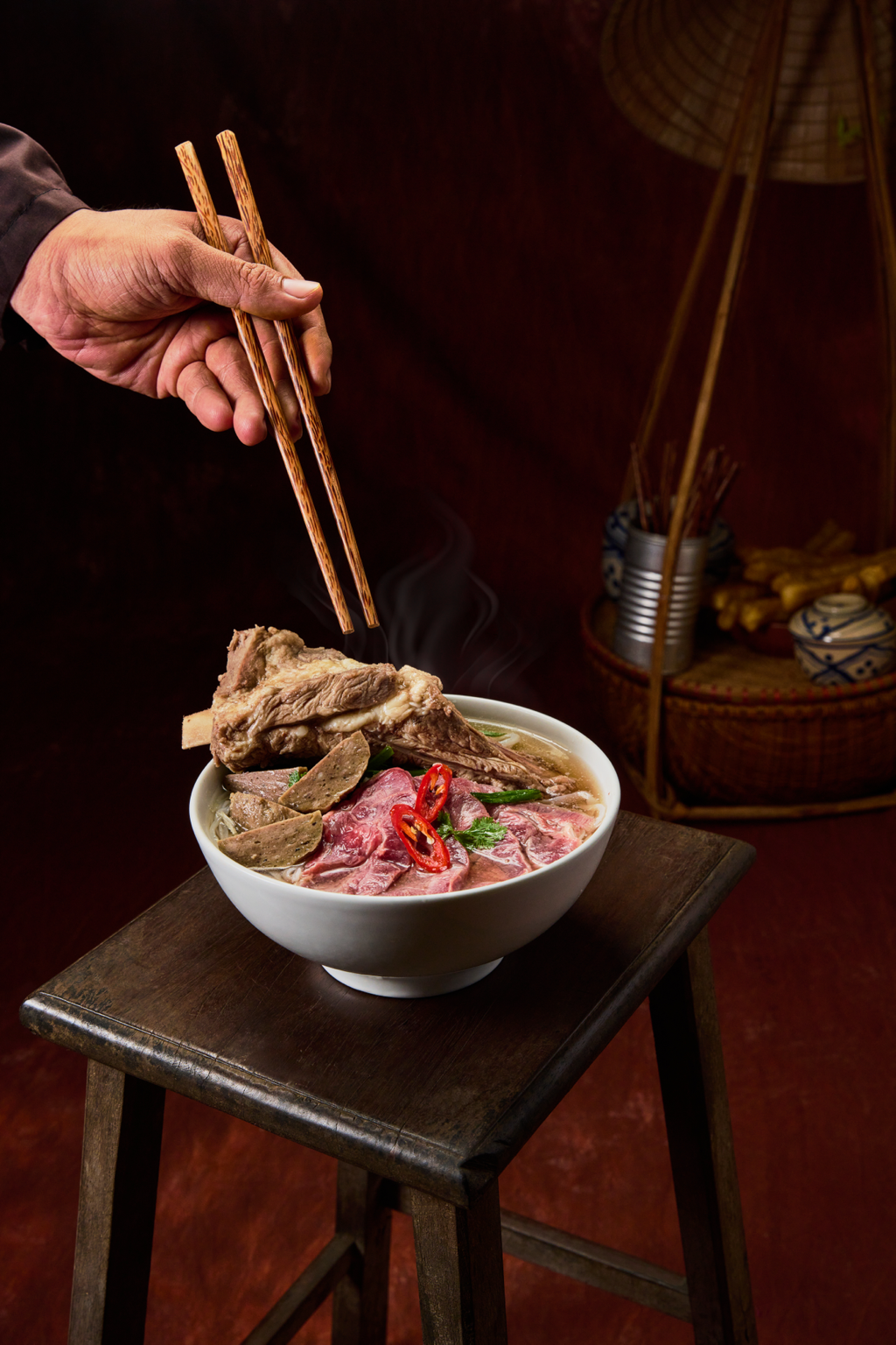 A person holding chopsticks above a bowl of steaming Asian-style beef Pho noodle soup, with slices of beef, herbs, and red chili peppers, on a small wooden table.