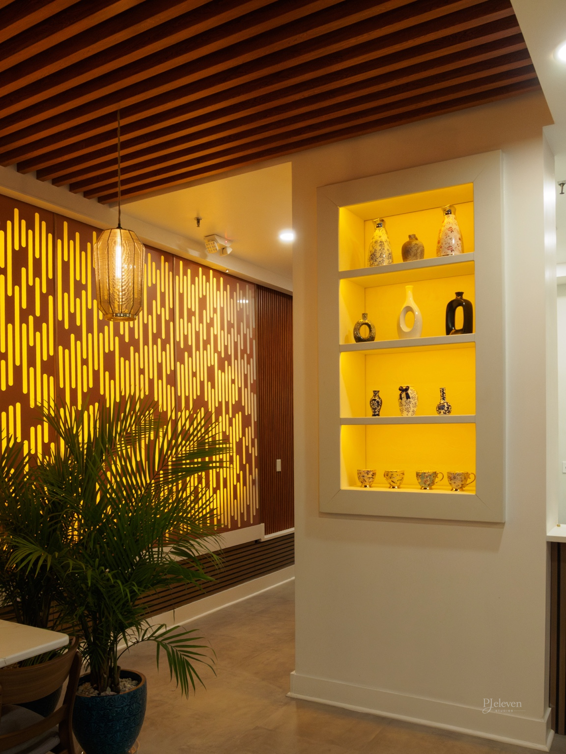 Interior view of a warmly lit room with a modern decorative yellow lighting feature, a wooden ceiling with a hanging lantern, a white wall with a built-in recessed display shelf filled with various vases and cups, and a potted plant in the foreground
