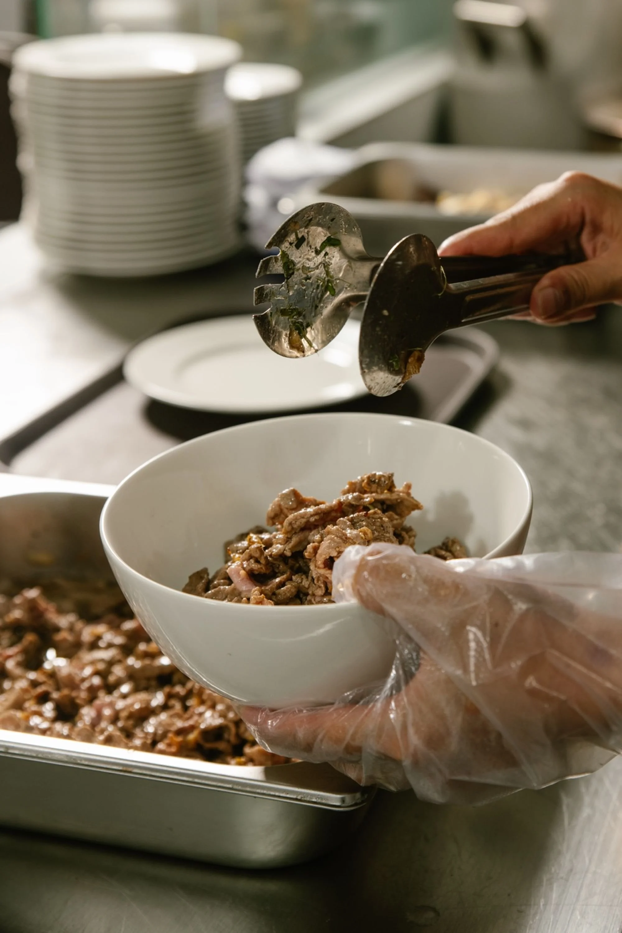 A person wearing a glove is filling a bowl with cooked, shredded ribeye beef from a serving tray, using metal tongs.