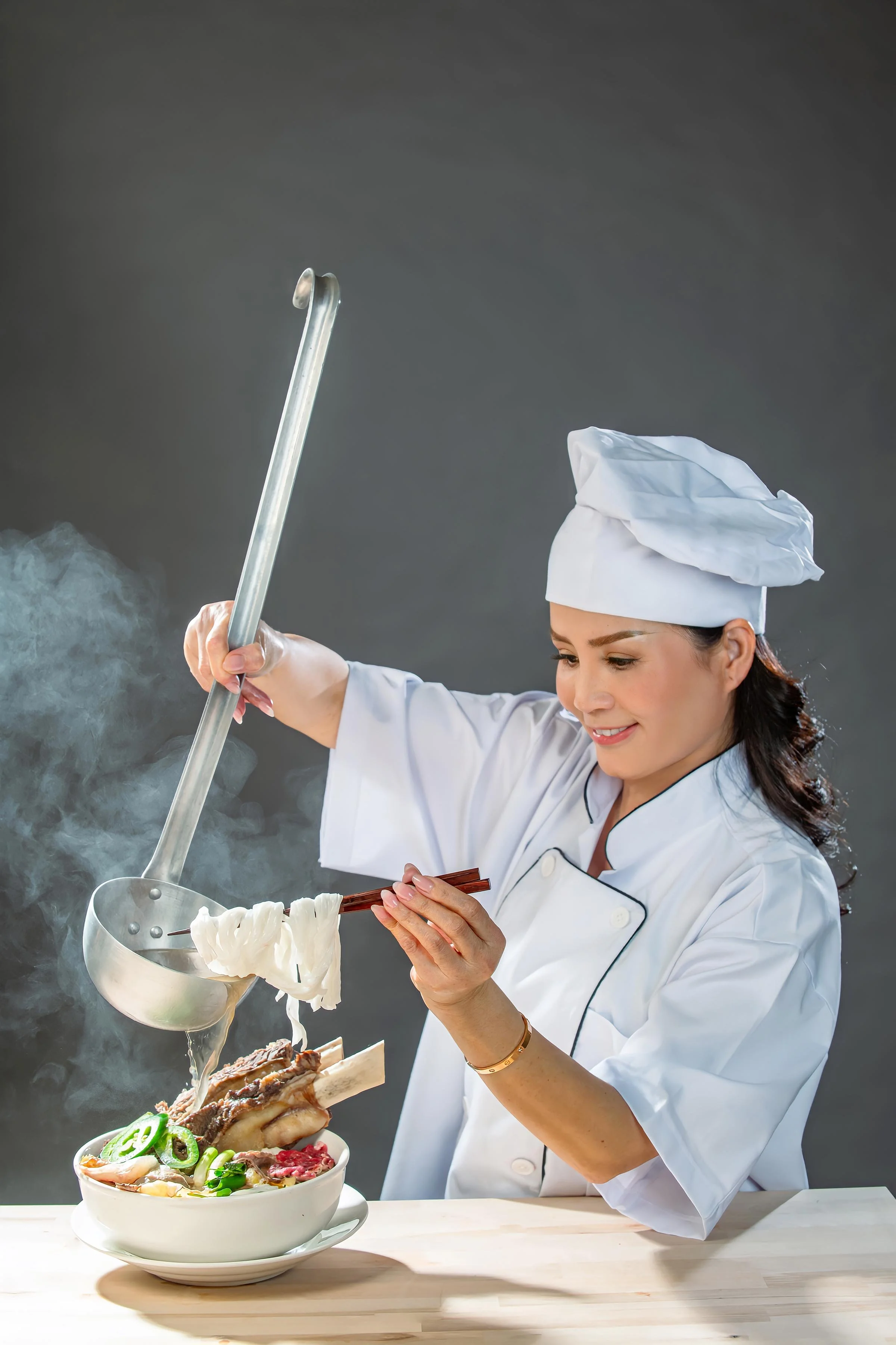 A female chef in a white uniform and chef hat is cooking noodles over a steaming bowl of Pho, which includes beef ribs, brisket, flank, green onions, and other ingredients, on a light wooden table against a dark background.