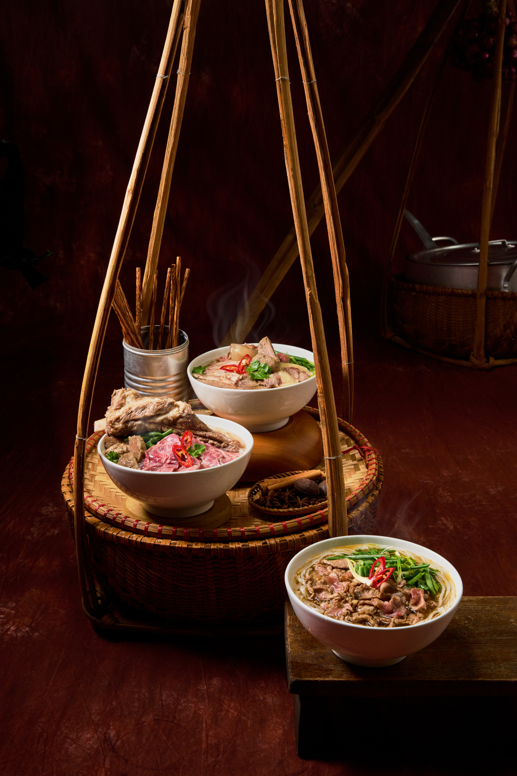 Four bowls of steaming Asian beef noodle soup on a wooden table, with a woven basket and chopsticks in the background.