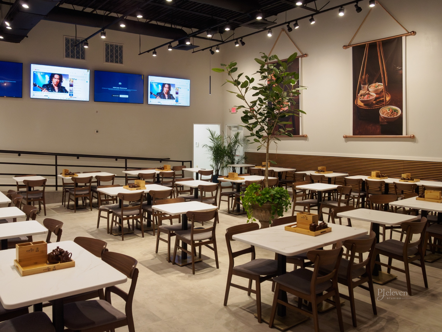 Empty restaurant dining area with white tables, brown chairs, potted plants, two large hanging banners with food images, multiple wall-mounted flat-screen TVs, and overhead lighting.