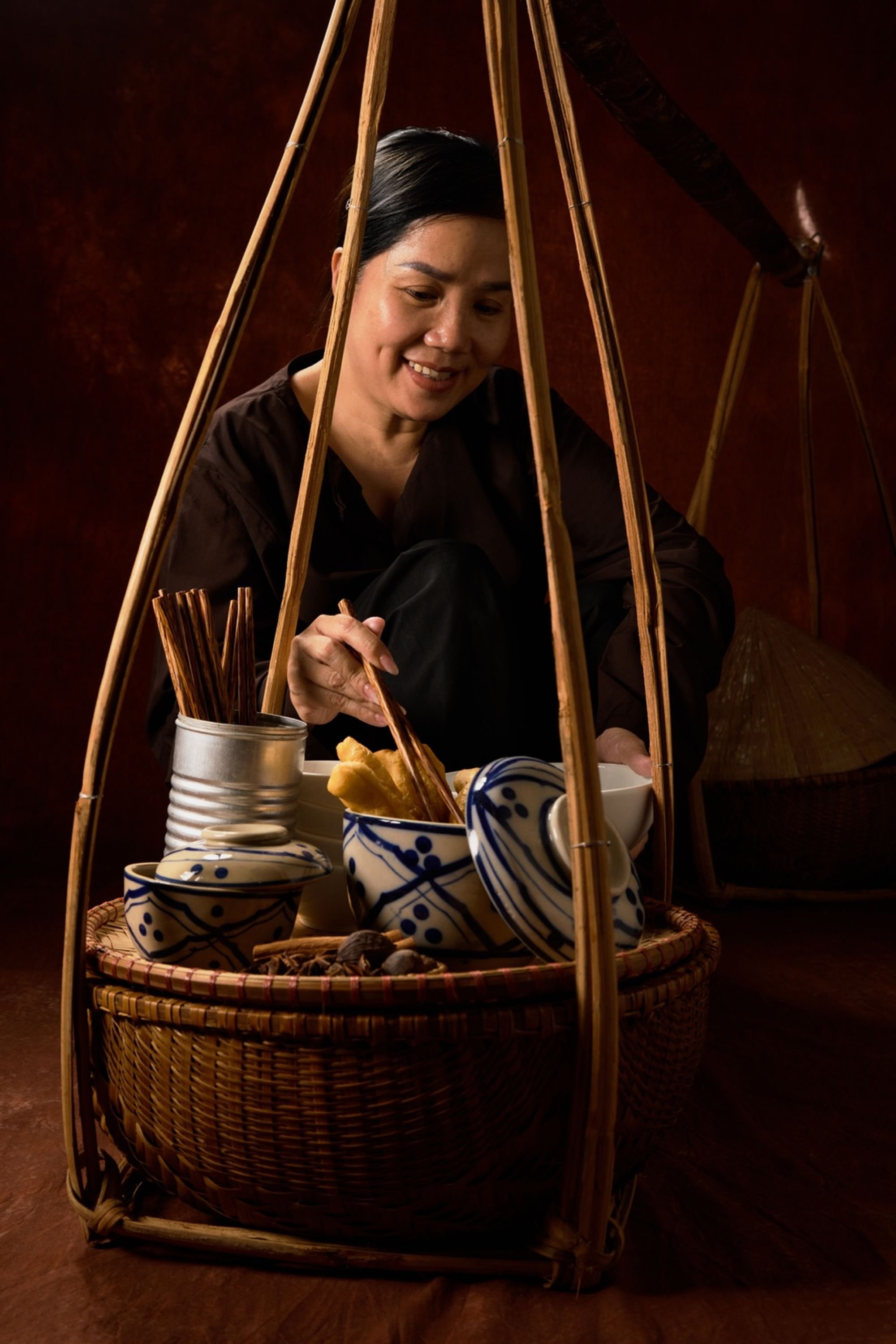 A woman is preparing traditional Japanese food using chopsticks, surrounded by ceramic bowls and utensils inside a decorative bamboo frame, with a dark background.