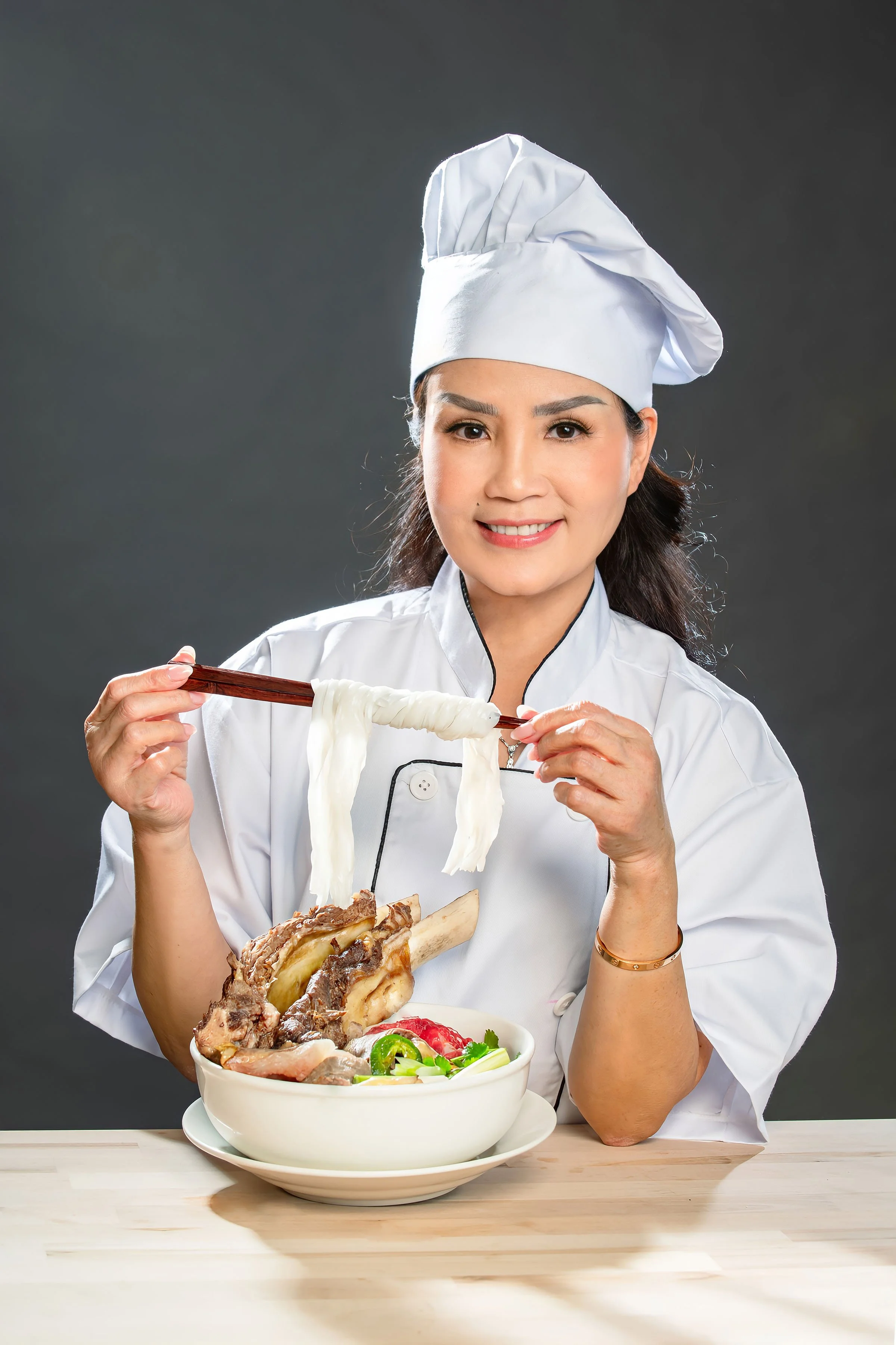 A female chef in a white uniform and hat holding chopsticks with noodles, sitting in front of Pho noodles bowl of hot soup with meat, vegetables, and noodles, against a dark background.