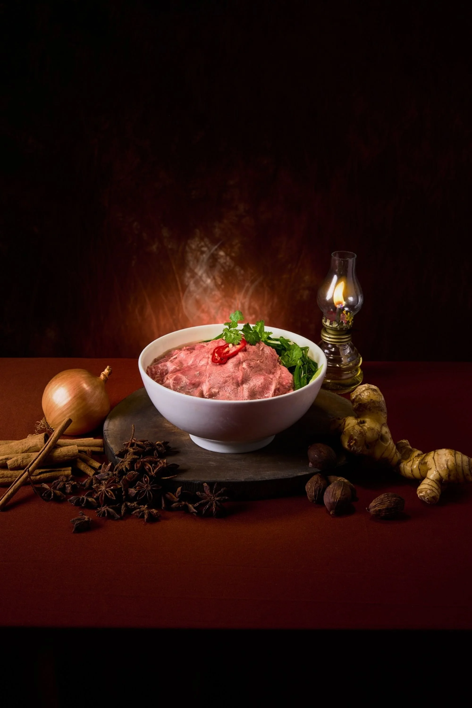 A bowl of raw ribeye beef surrounded by spices and fresh ingredients on a dark wooden surface, illuminated by a small oil lamp.