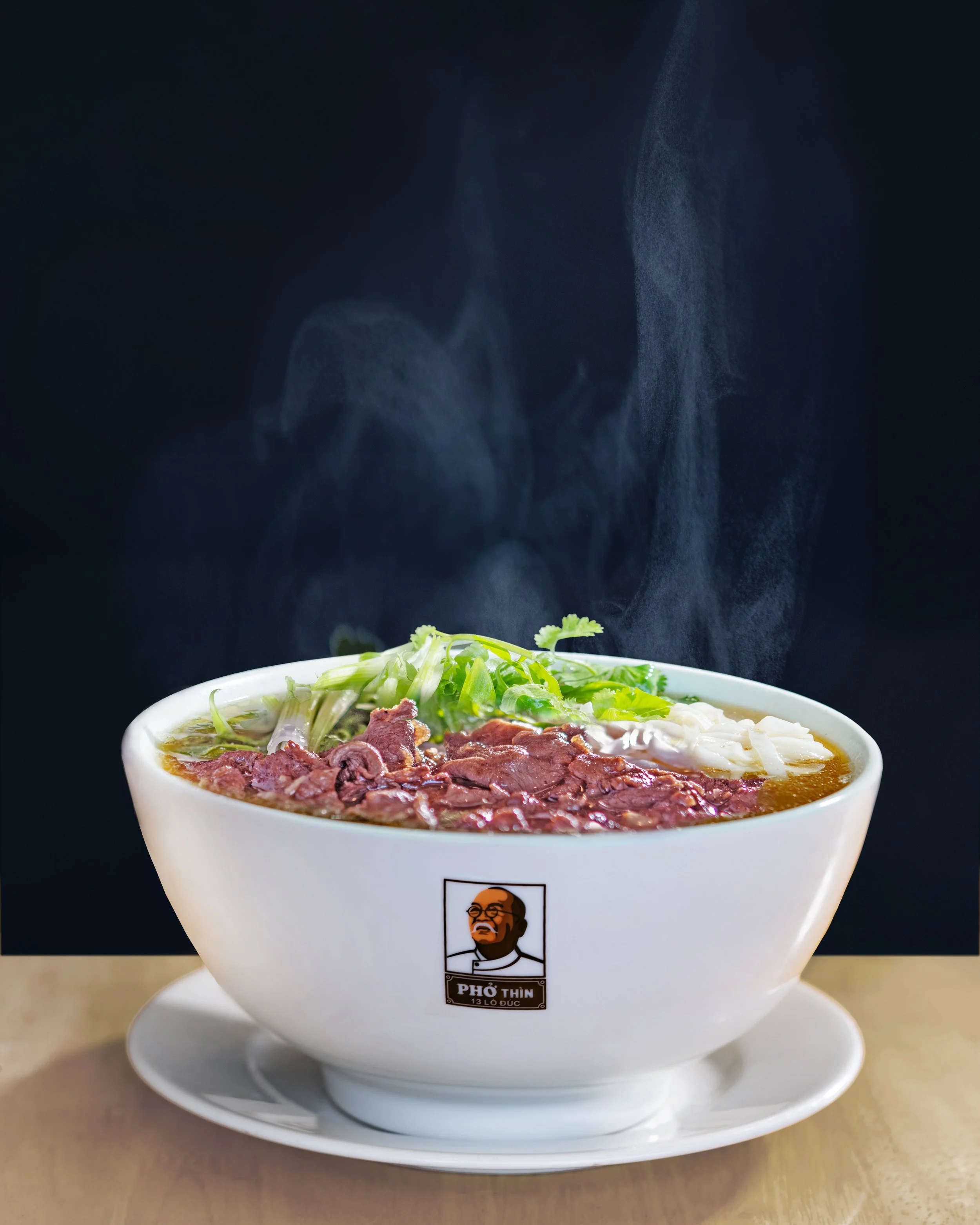 A steaming bowl of beef pho with sliced beef, green onions, and rice noodles in a white bowl with a logo, placed on a white saucer on a wooden surface.