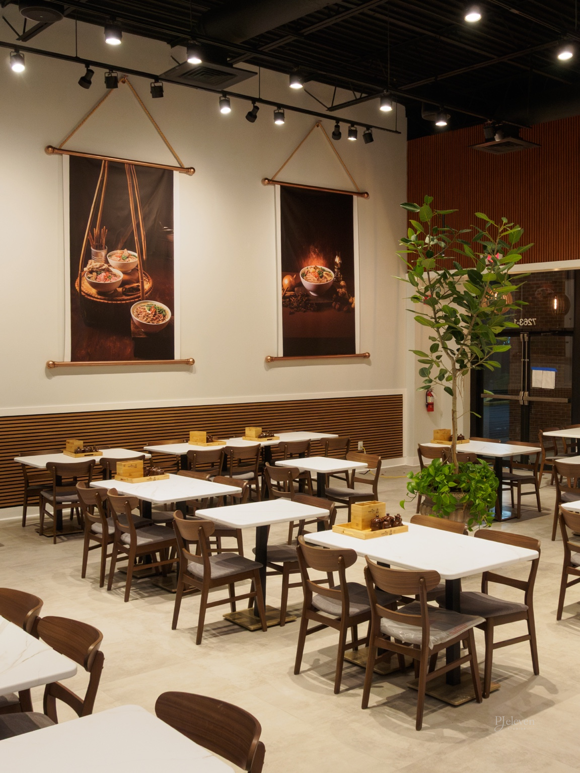 Empty restaurant dining area with white tables, wooden chairs, large green plant, and wall art featuring Asian Vietnamese cuisine.