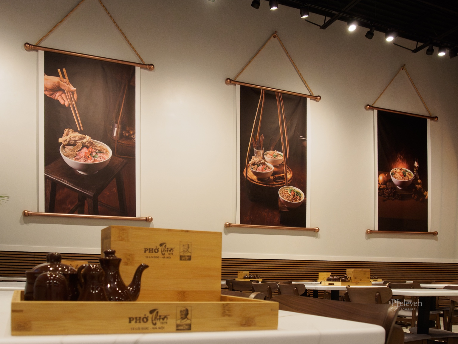 Interior of a restaurant dining area with Asian-themed wall art featuring bowls of Pho noodles, hanging banners with images of Asian dishes, wooden tables, and chairs, and a wooden condiment tray with teapots in the foreground.