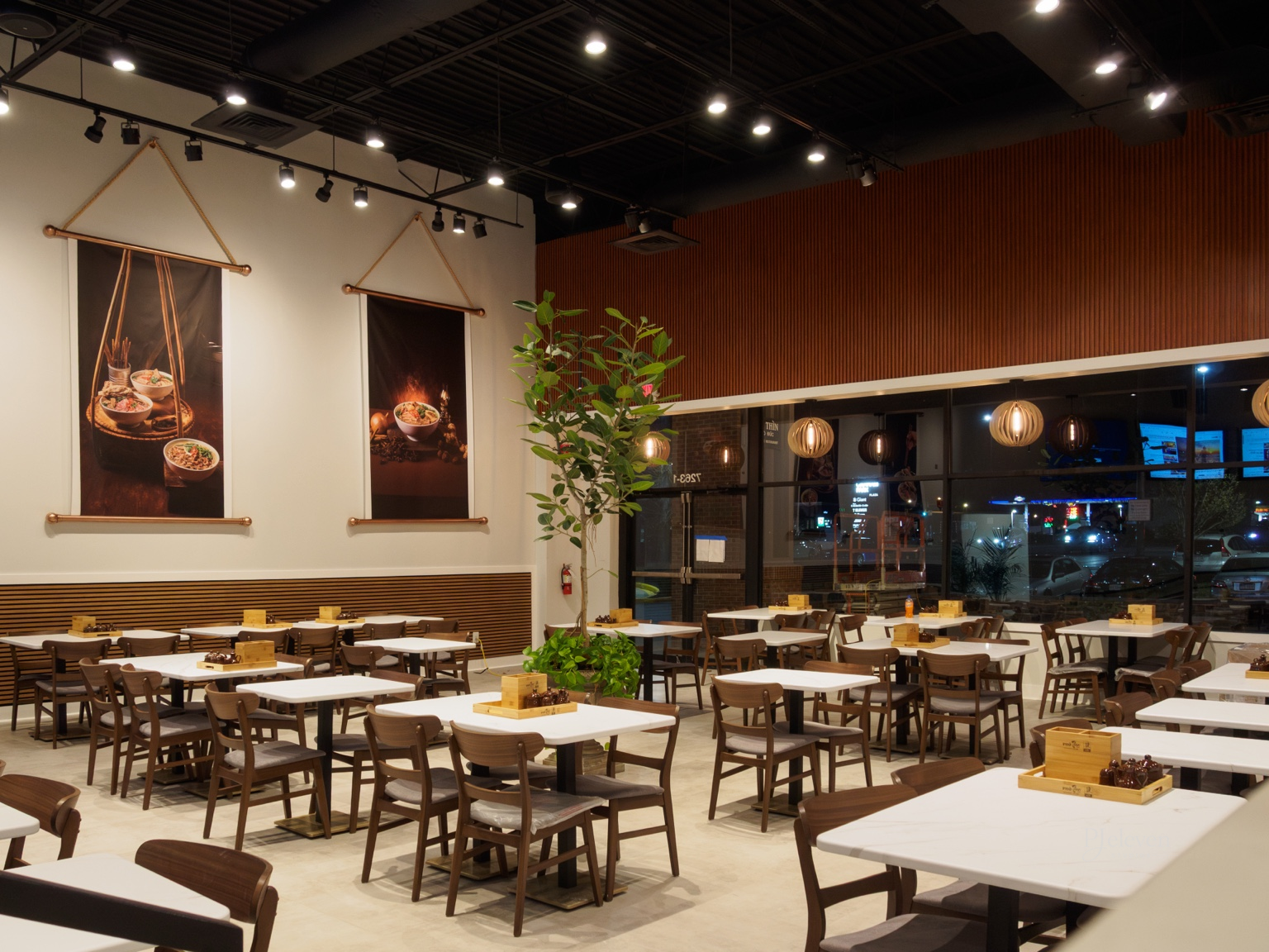 Empty restaurant with white tables, wooden chairs, potted plant, and hanging lights. Night view outside the large glass windows.