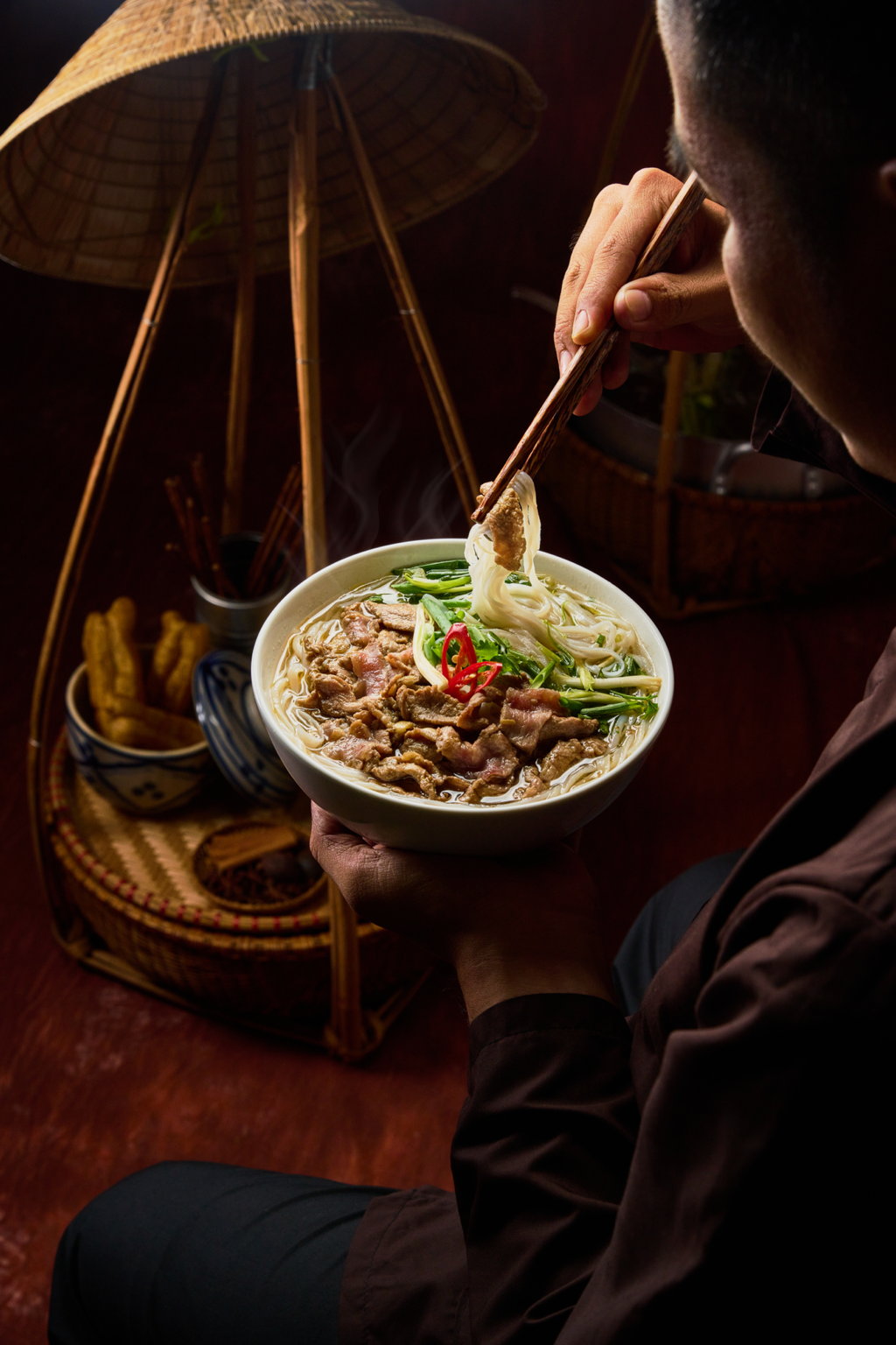 Person eating bowl of noodle soup with beef, vegetables, green onion and red chili slices, using chopsticks, in a cozy setting.