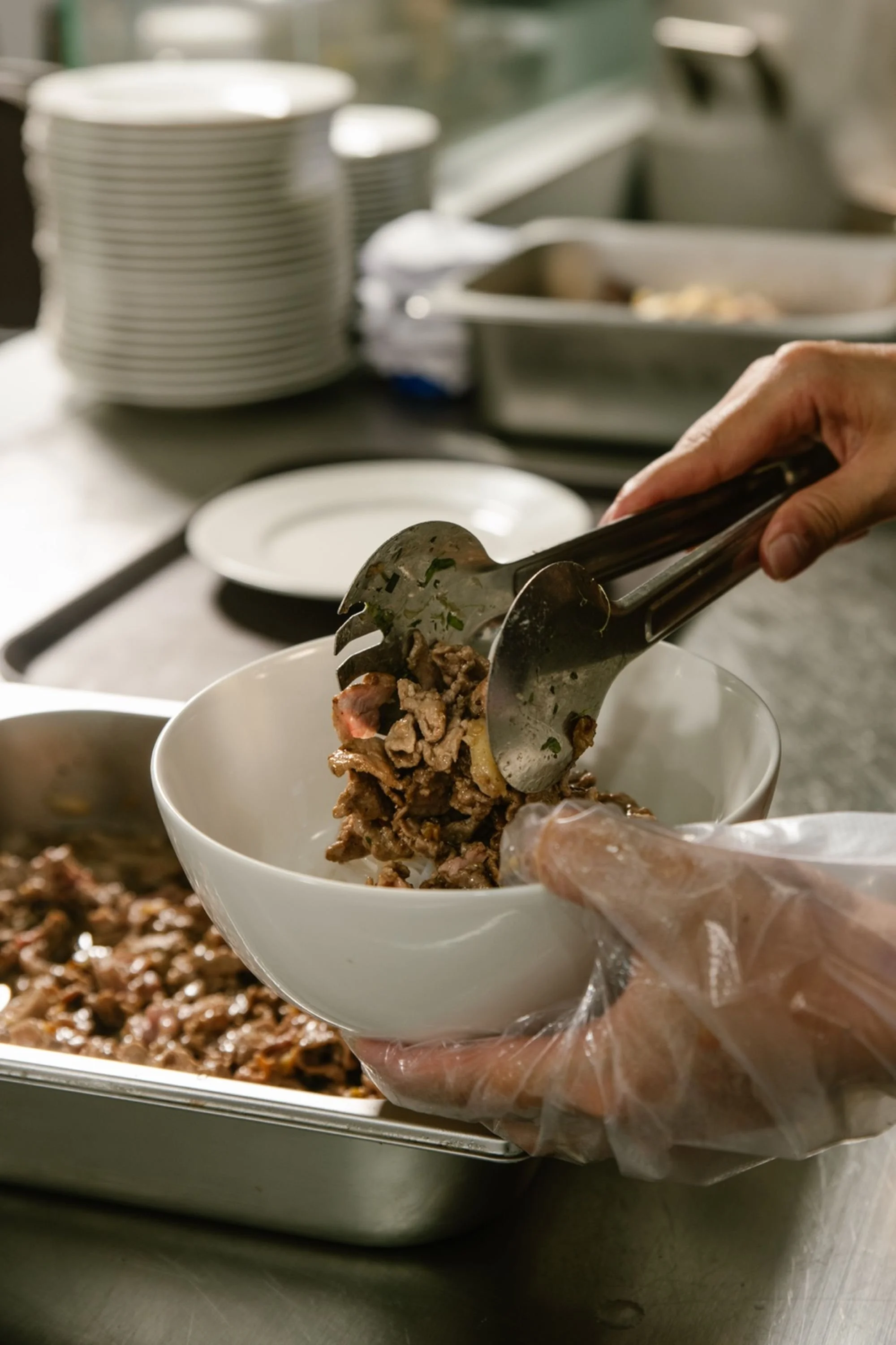 Person serving cooked ribeye meat into a white bowl at a food service station, with stacked plates and kitchen supplies in the background.