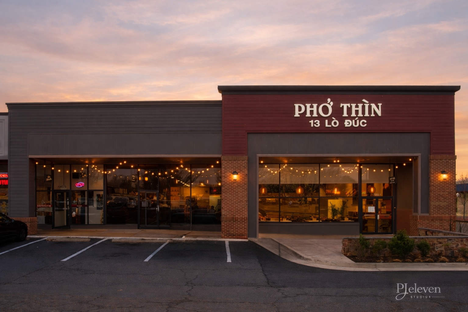 Exterior of a Vietnamese restaurant called Pho Thin at 13 Lò Đúc, with large glass windows, string lights, and a parking lot in front at sunset.