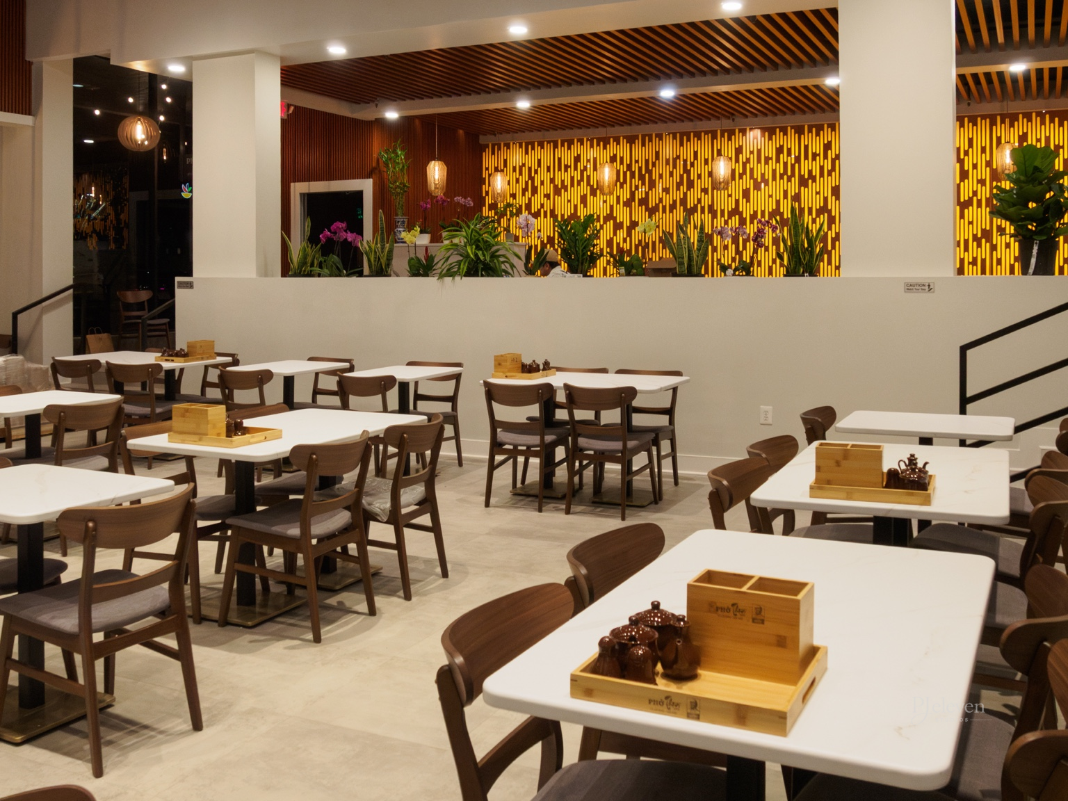 Interior of a restaurant with white tables, wooden chairs, and decorative plants, featuring warm lighting and a patterned yellow and black accent wall.