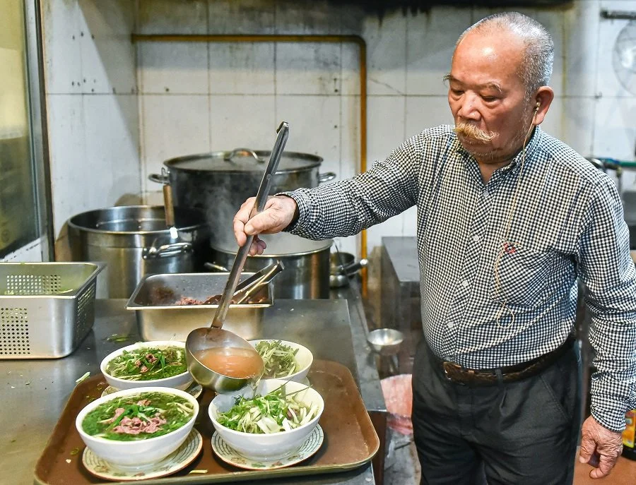 An elderly man serving soup and preparing a bowl of hot Pho beef noodles in a kitchen setting.