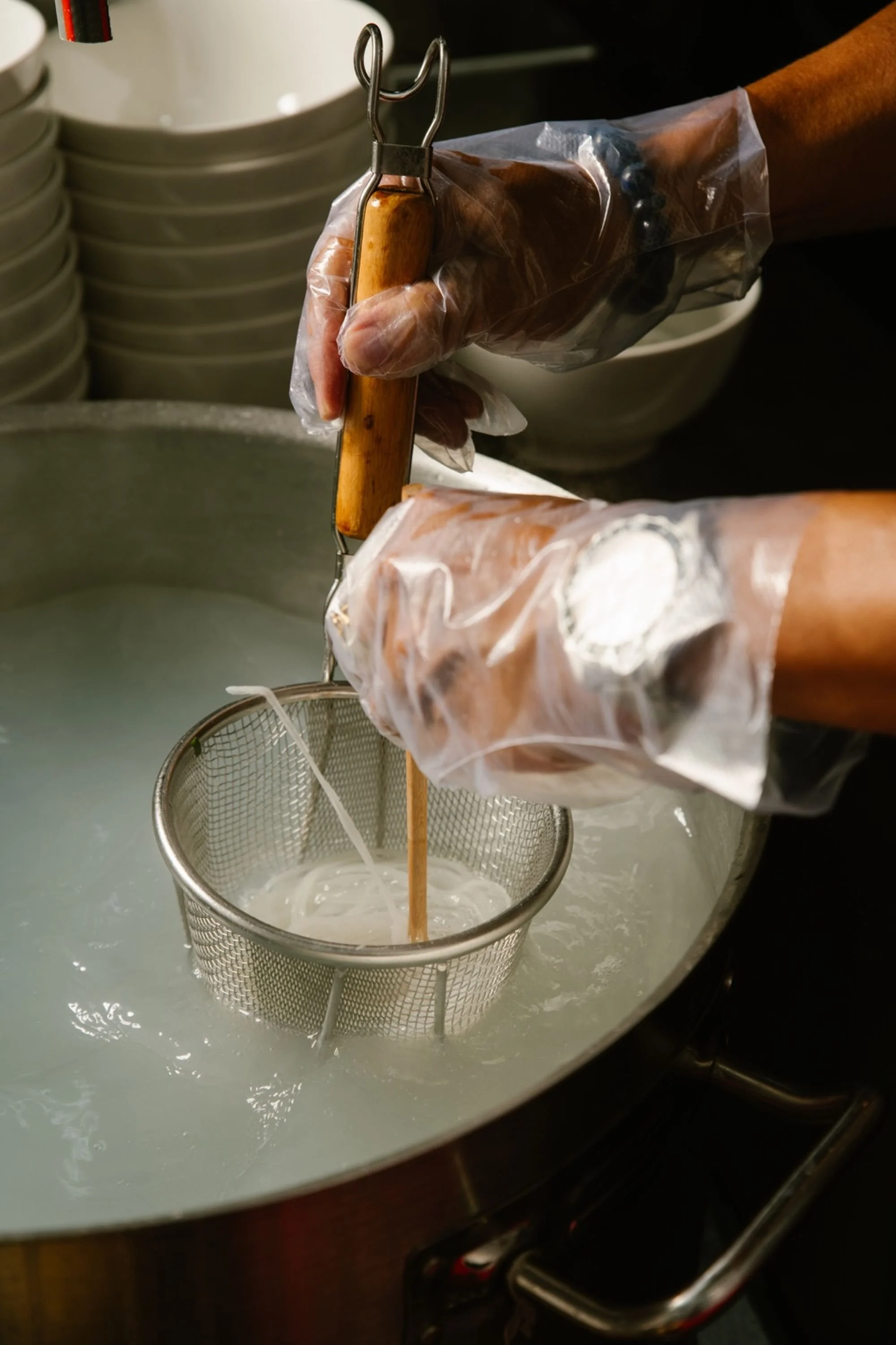 Person wearing plastic gloves is using a slotted spoon to strain hot liquid into a metal mesh strainer over a large pot of boiling water and fresh rice noodles.