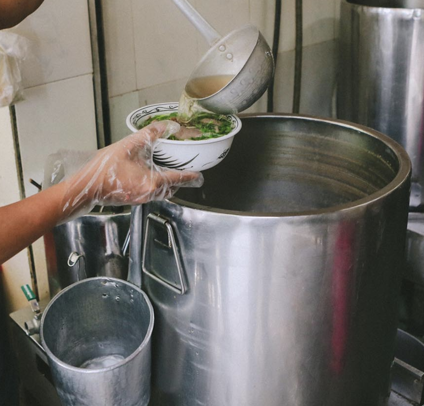 A person wearing a plastic glove is pouring soup from a metal ladle into a bowl, with a large stainless steel pot and kitchen tiles in the background.