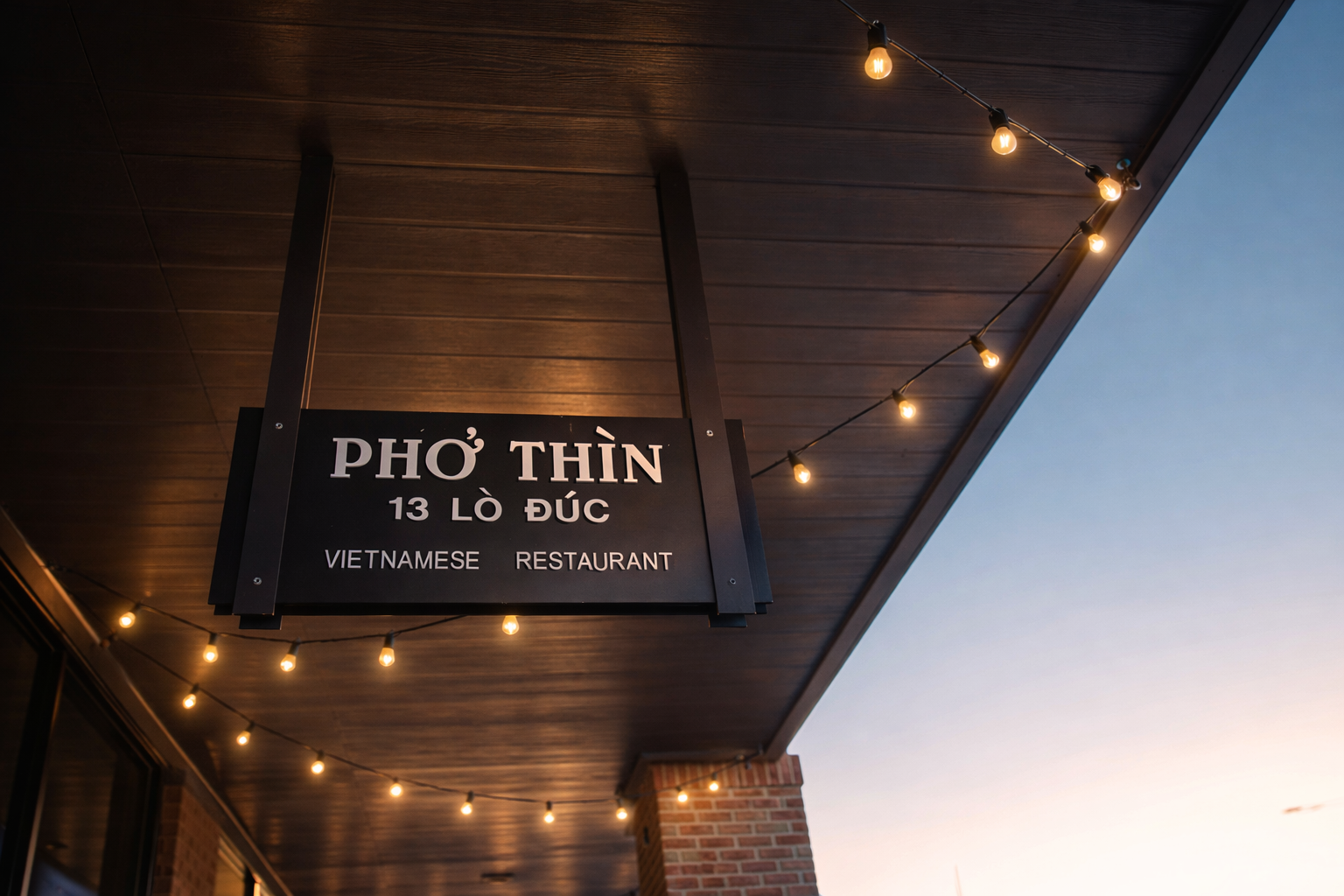 Sign for Pho Thin Vietnamese restaurant with string lights against a wooden ceiling and brick wall in the evening.