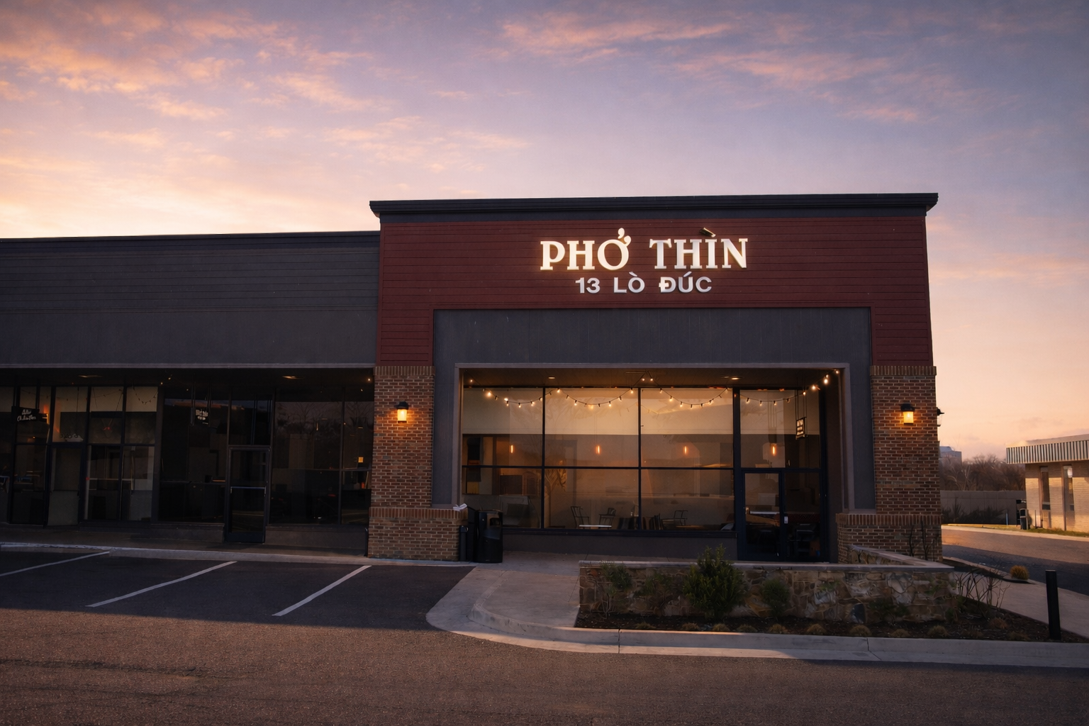 Exterior view of Pho Thin restaurant during sunset, with illuminated sign, large windows, and parking lot in front.