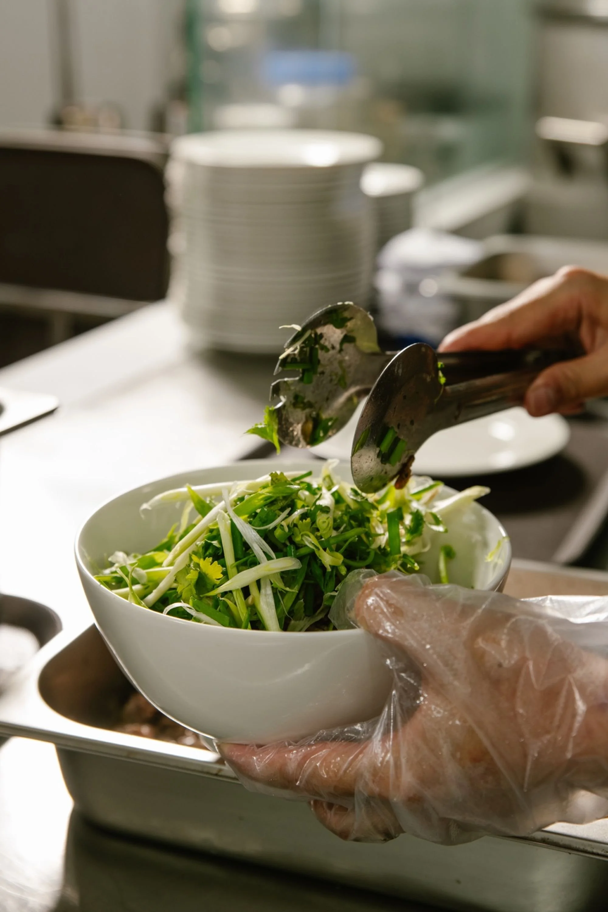 A person wearing a plastic glove is adding fresh green onions and vegetables into a white bowl with tongs, in a kitchen or food preparation setting.