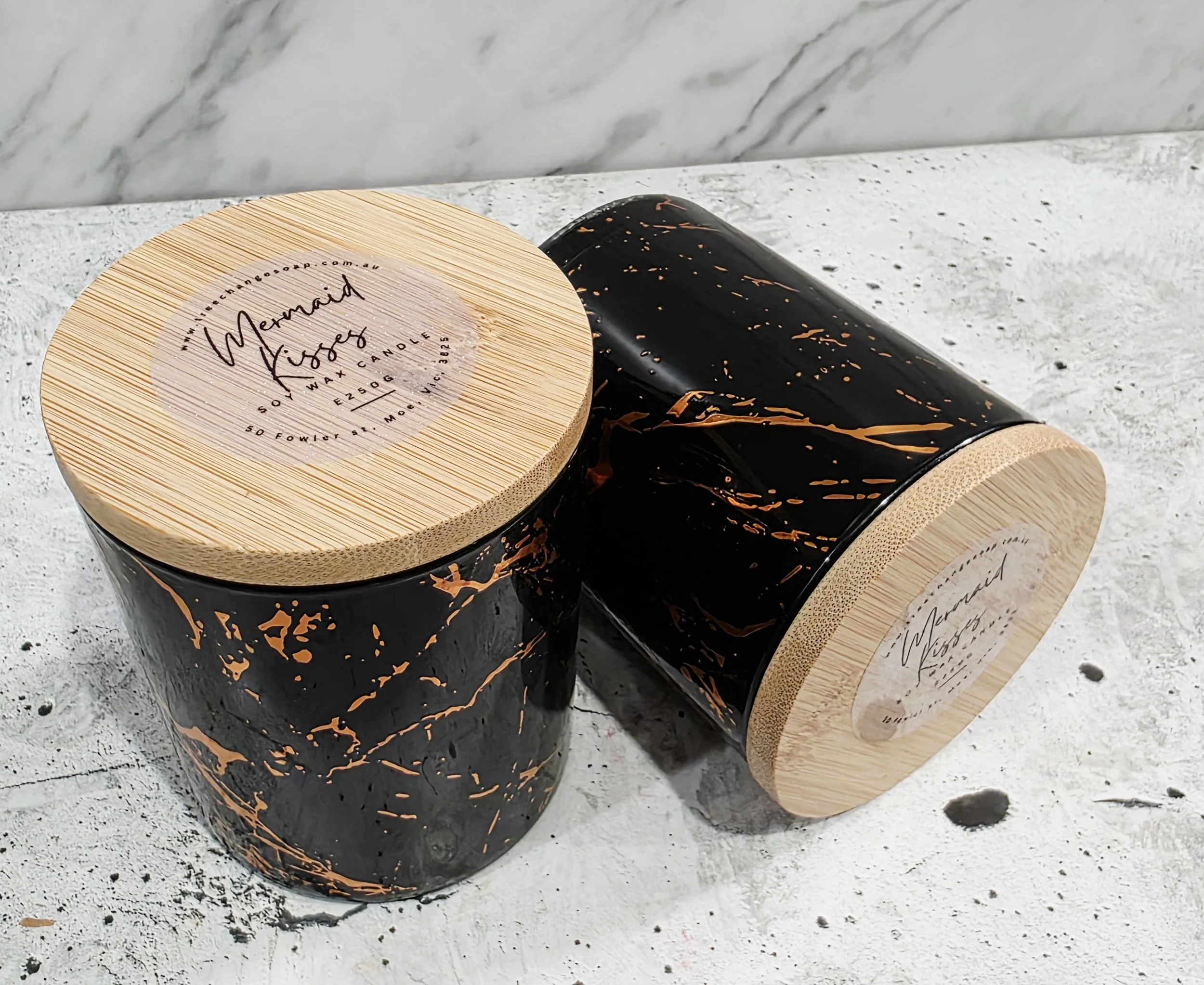 Two black and gold marble scented soy wax candles with wooden lids, placed on a textured gray surface.