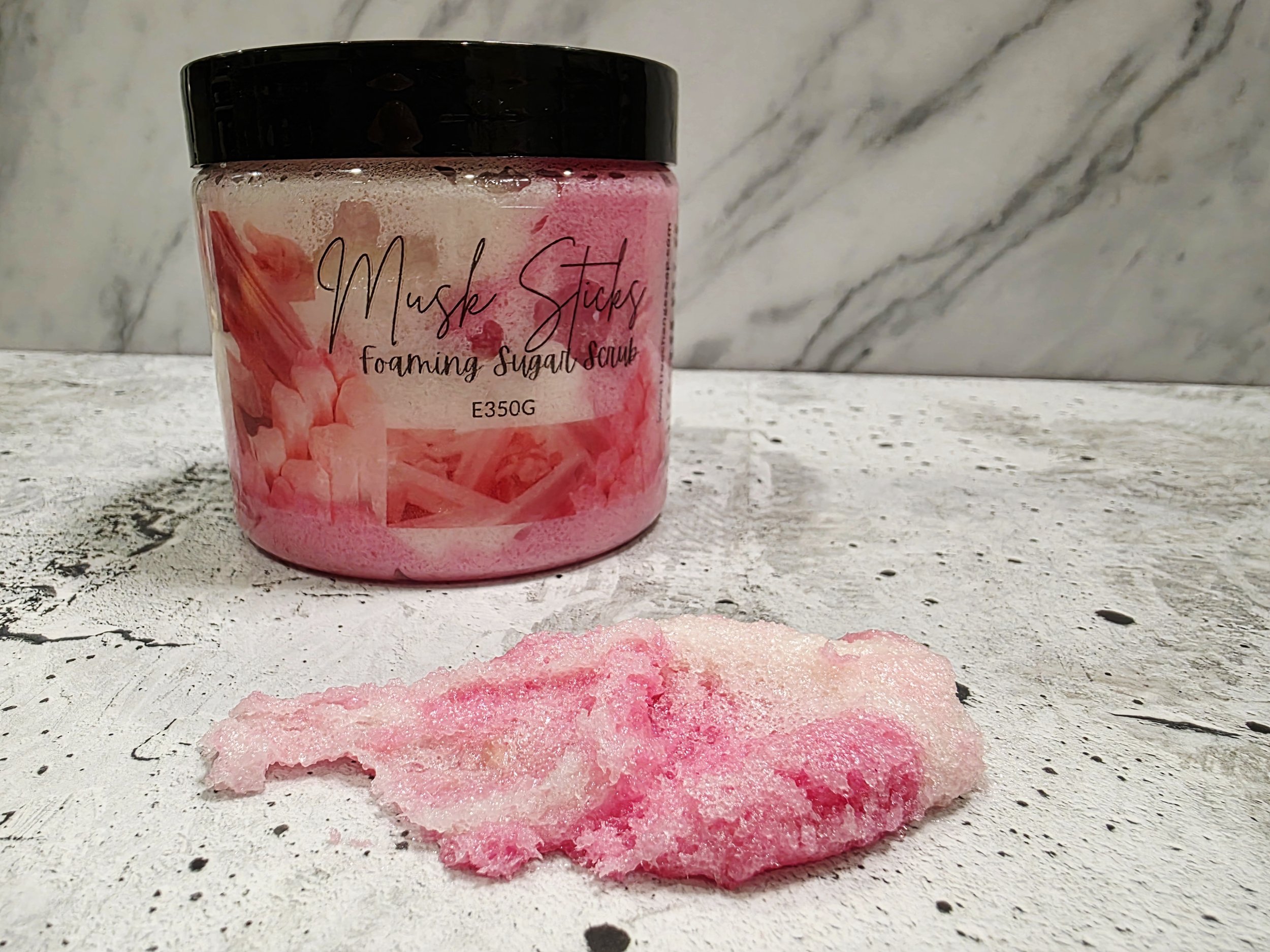 A pink frosted container of Musk Sticks foaming sugar scrub on a white textured surface with a pink and white scrub in front of it.