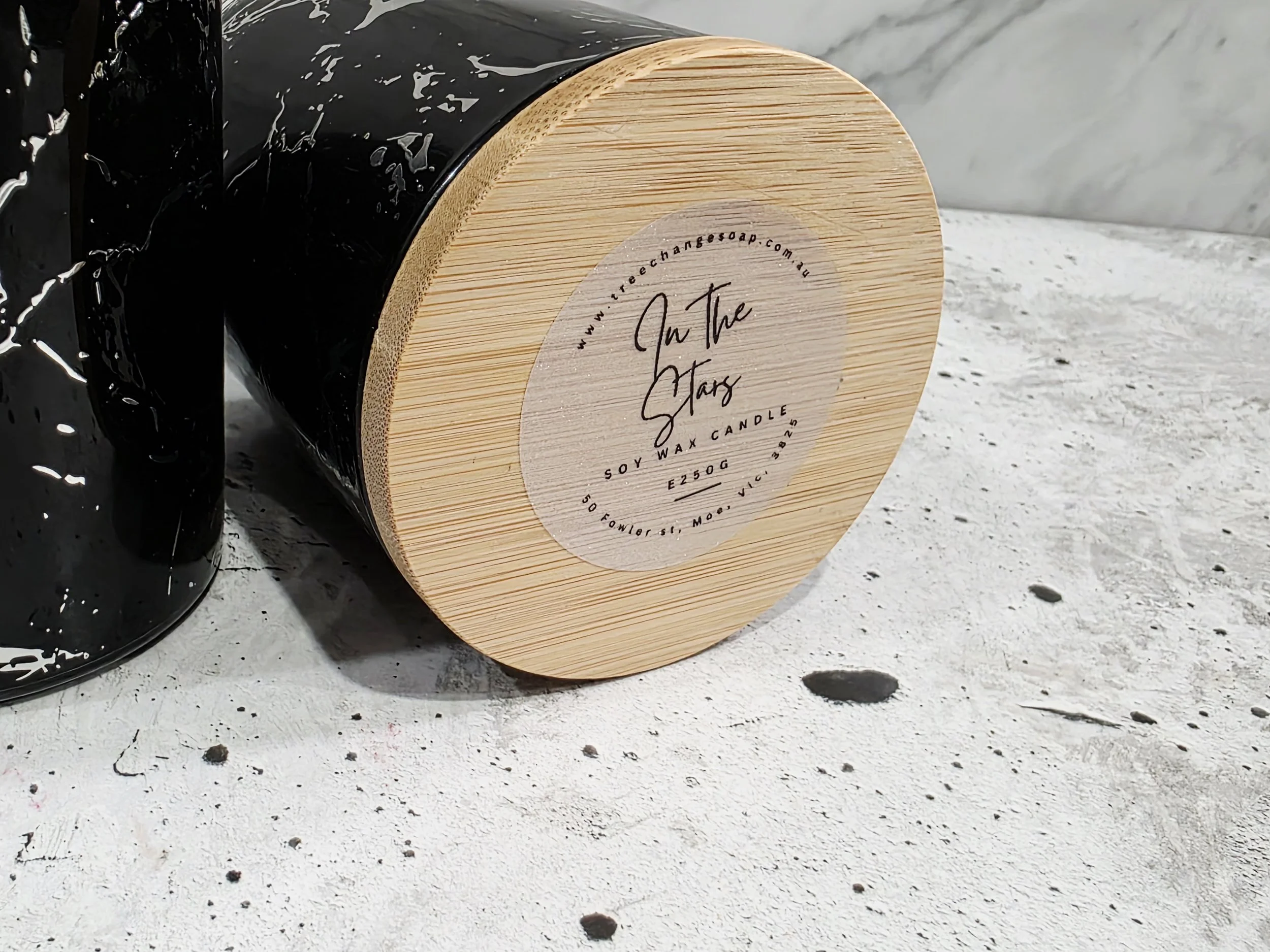 A black soy wax candle in a black jar and a round wooden lid with a label that reads 'On The Stars' placed on a textured white surface.