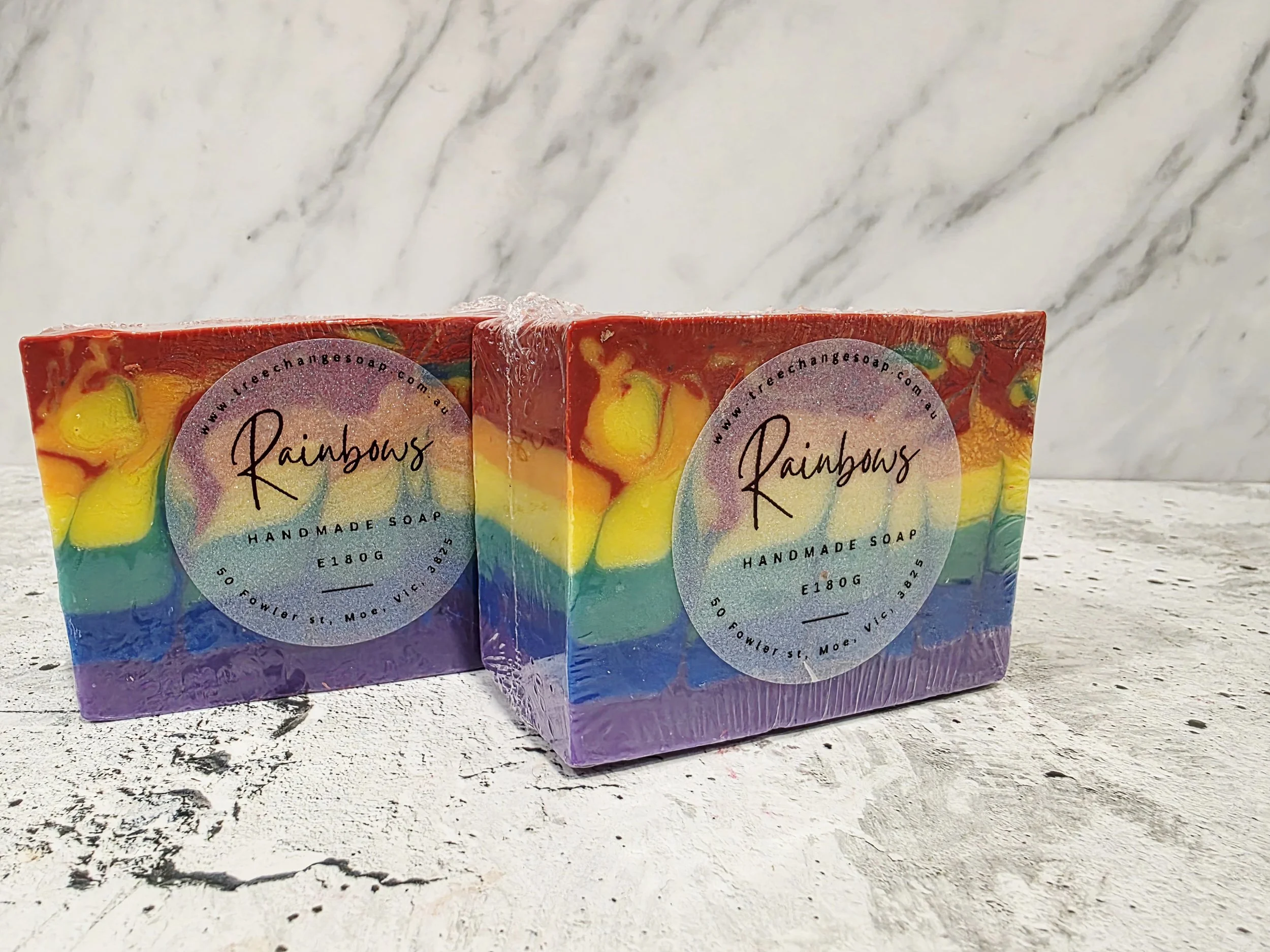 Two bars of rainbow-colored handmade soap with a label that says 'Rainbows, handmade soap, 180g' on a textured white surface with a marble background.