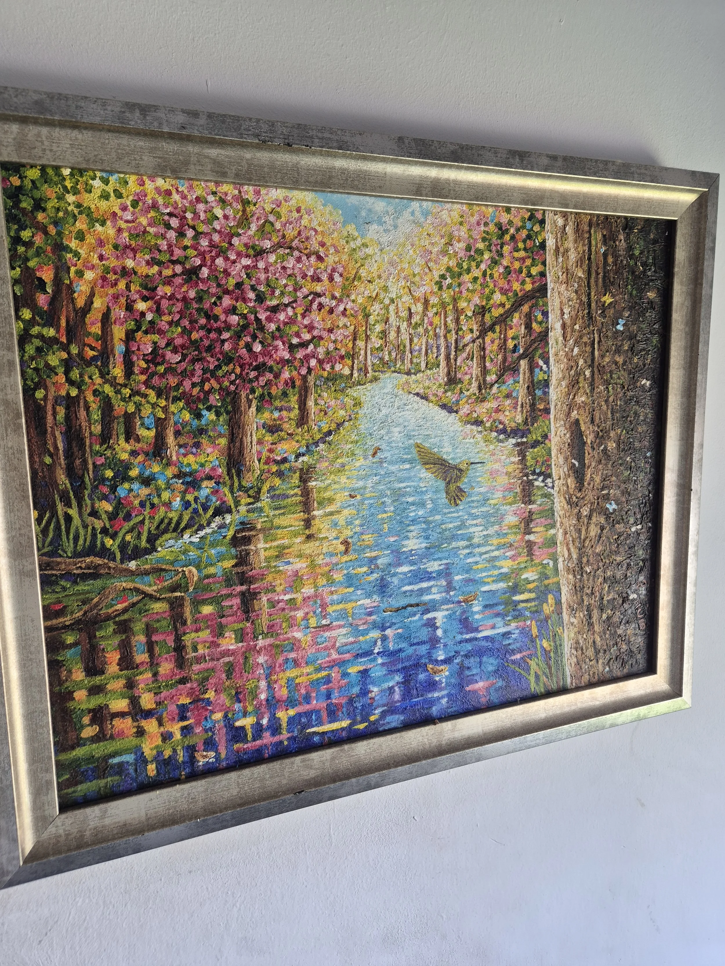 A framed oil painting depicting a vibrant, colourful  forest reflecting in a calm river, with a single bird flying low over the waters surface.