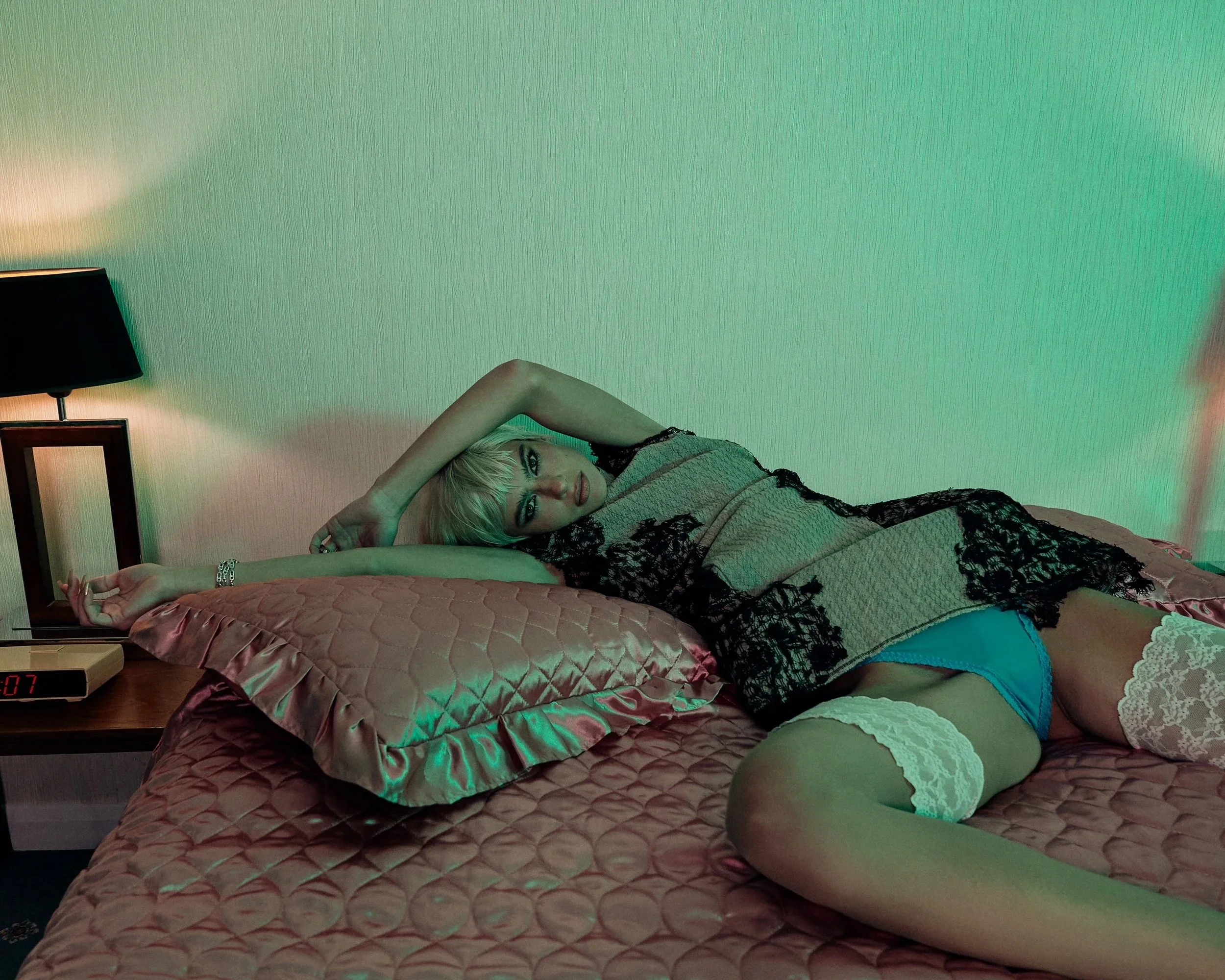 A woman lying on a bed with pink bedding and lace lingerie, looking at the camera with a relaxed pose.