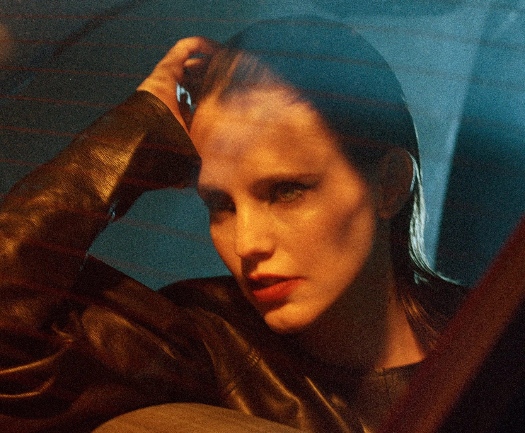Anna Calvi with wet hair and intense makeup, wearing a shiny brown leather jacket, posing with her hand on her head against a dark blue background.