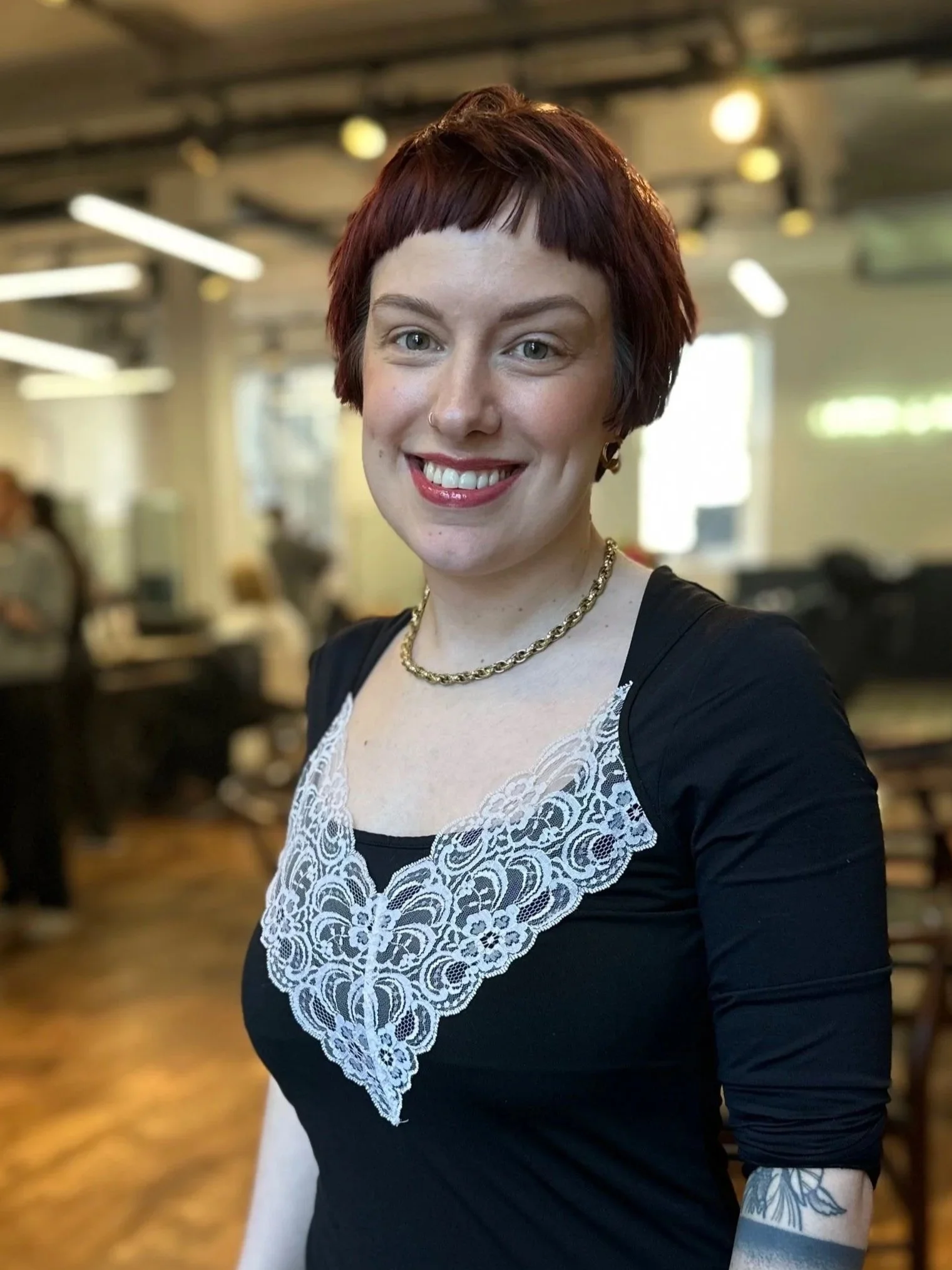A woman with short red hair, smiling, wearing a black dress with lace detailing on the front and a gold chain necklace. Inside a room with wood flooring and blurred background of people and furniture.