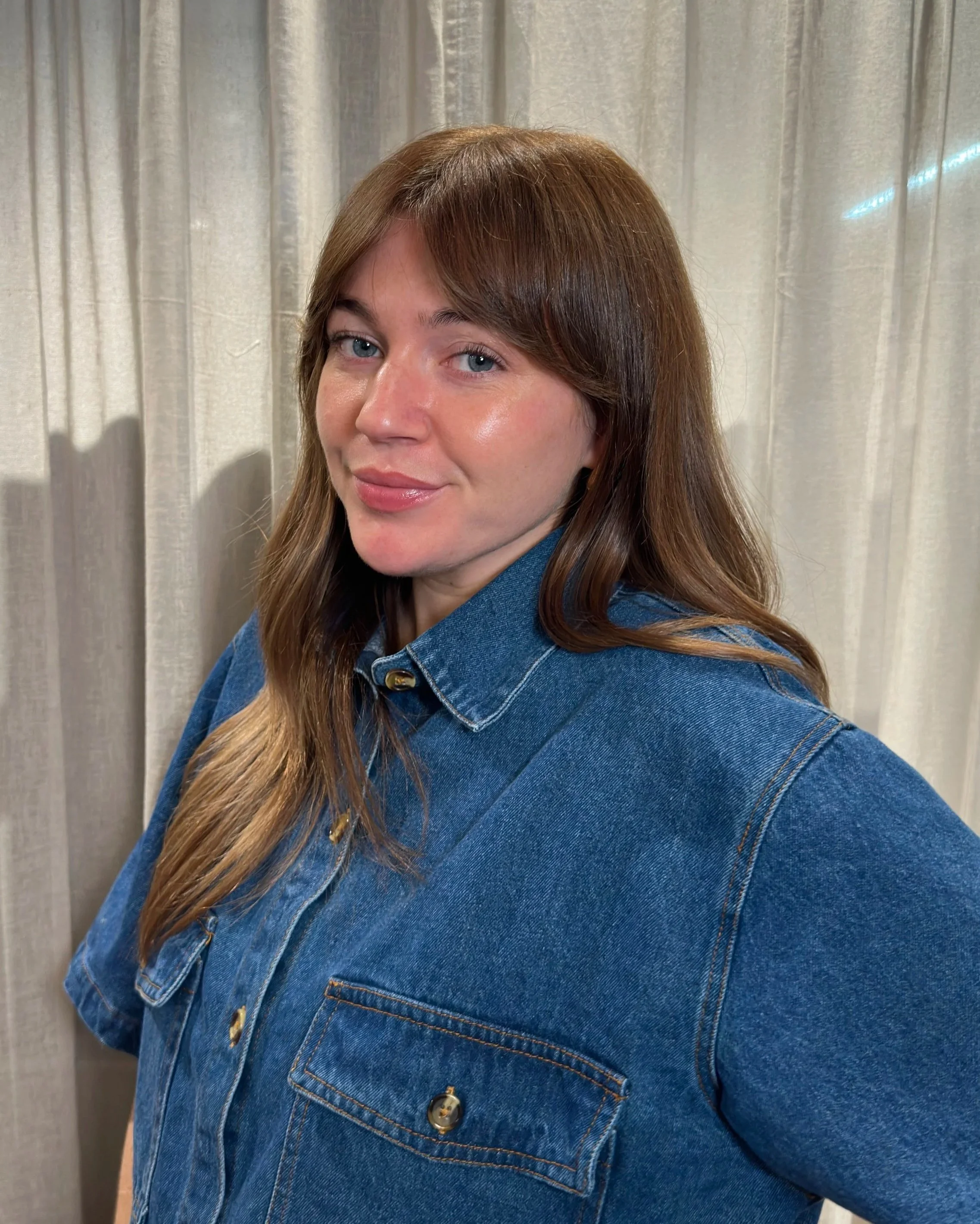 A woman with long brown hair, wearing a black and white striped shirt and black pants, leaning against a wall in a room with a mirror and chairs in the background.