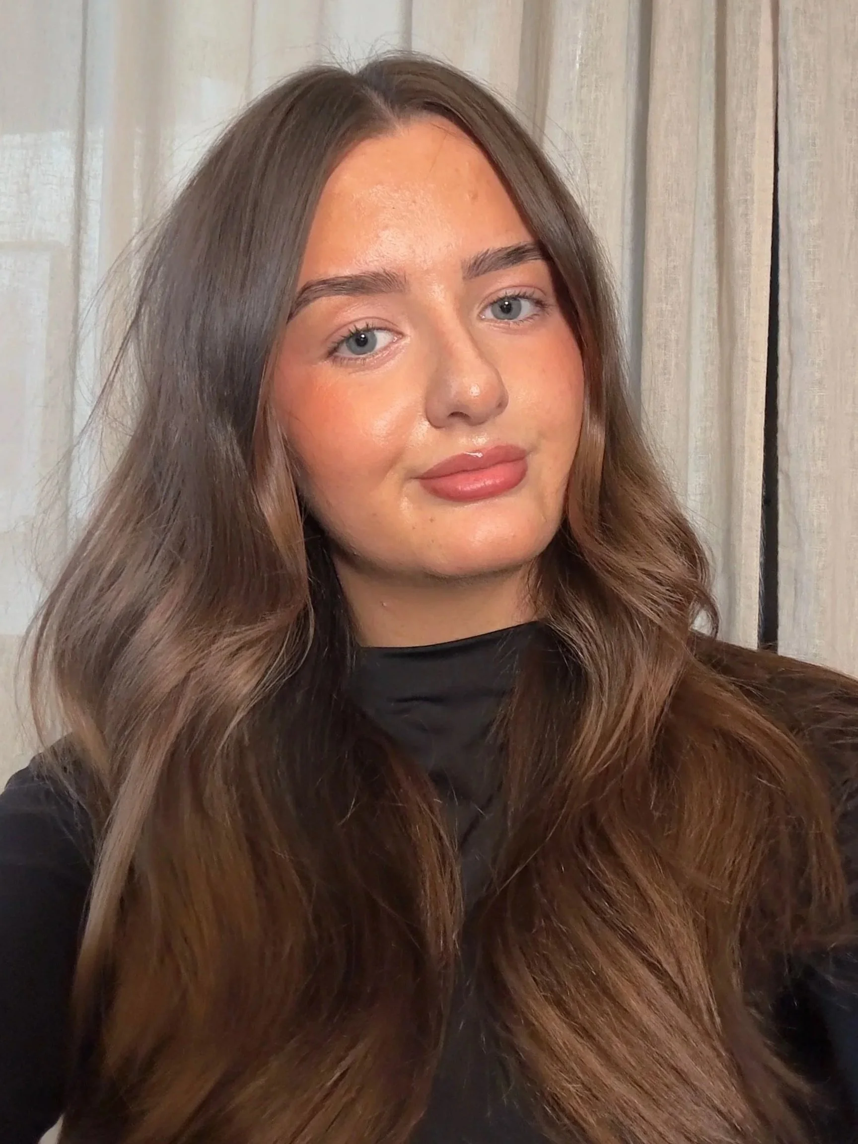 A woman with long, wavy brown hair and fair skin, wearing a black top, standing in front of beige curtains.